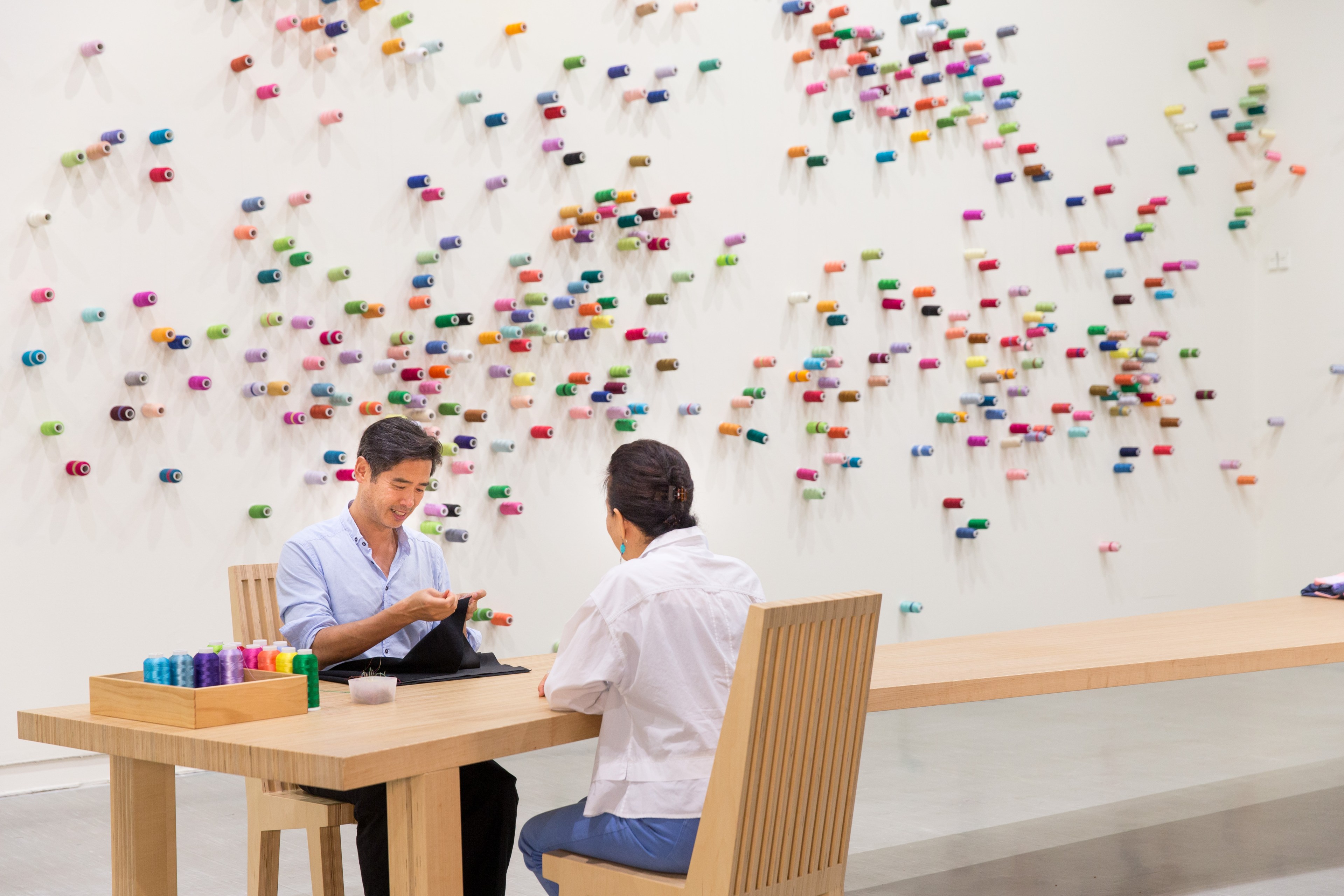 Two people sit at a table in an art gallery with colorful spools on the wall behind them.