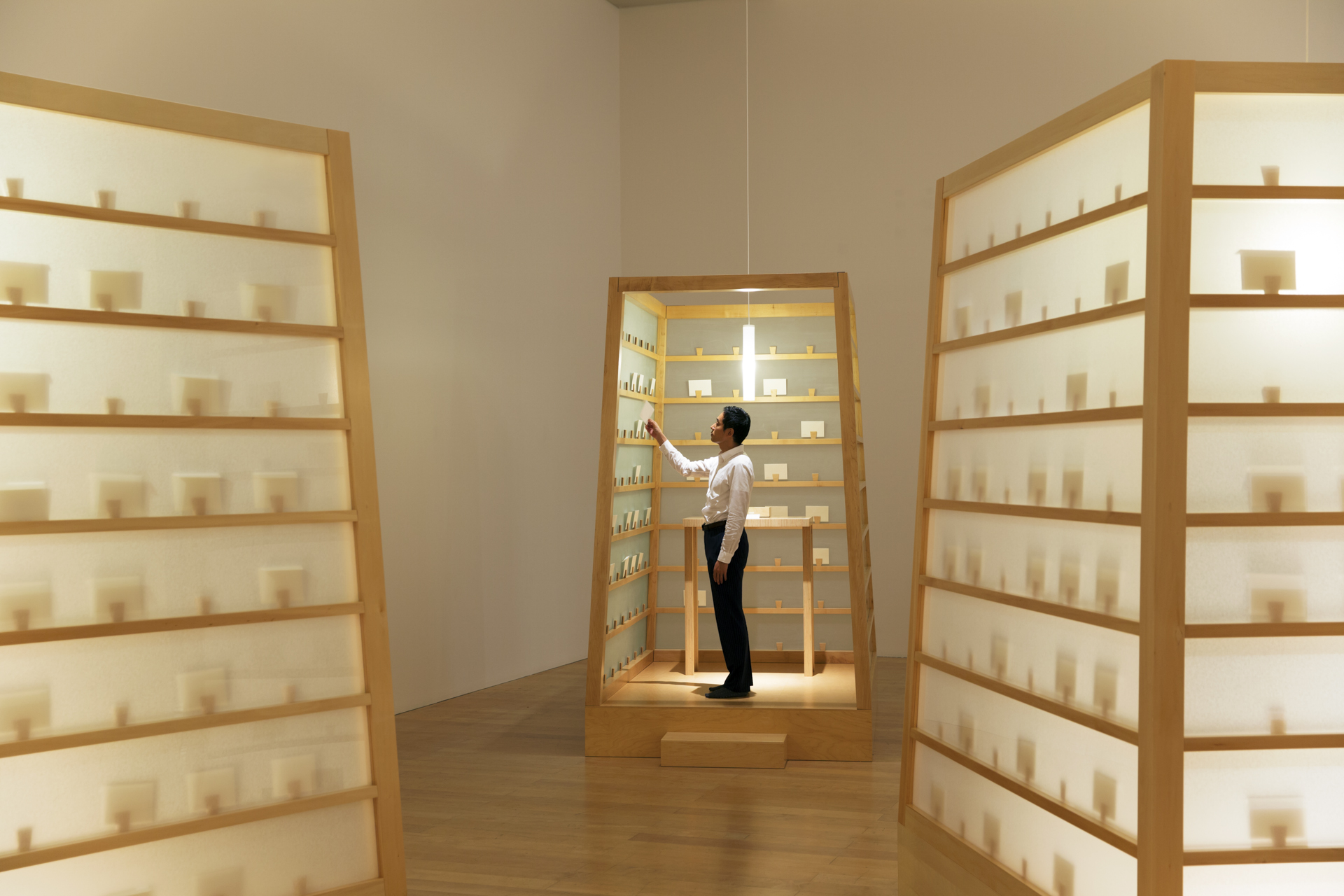 A person stands inside a lit, box-like wooden shelf structure, reaching out to a small object amidst many similar objects on shelves.