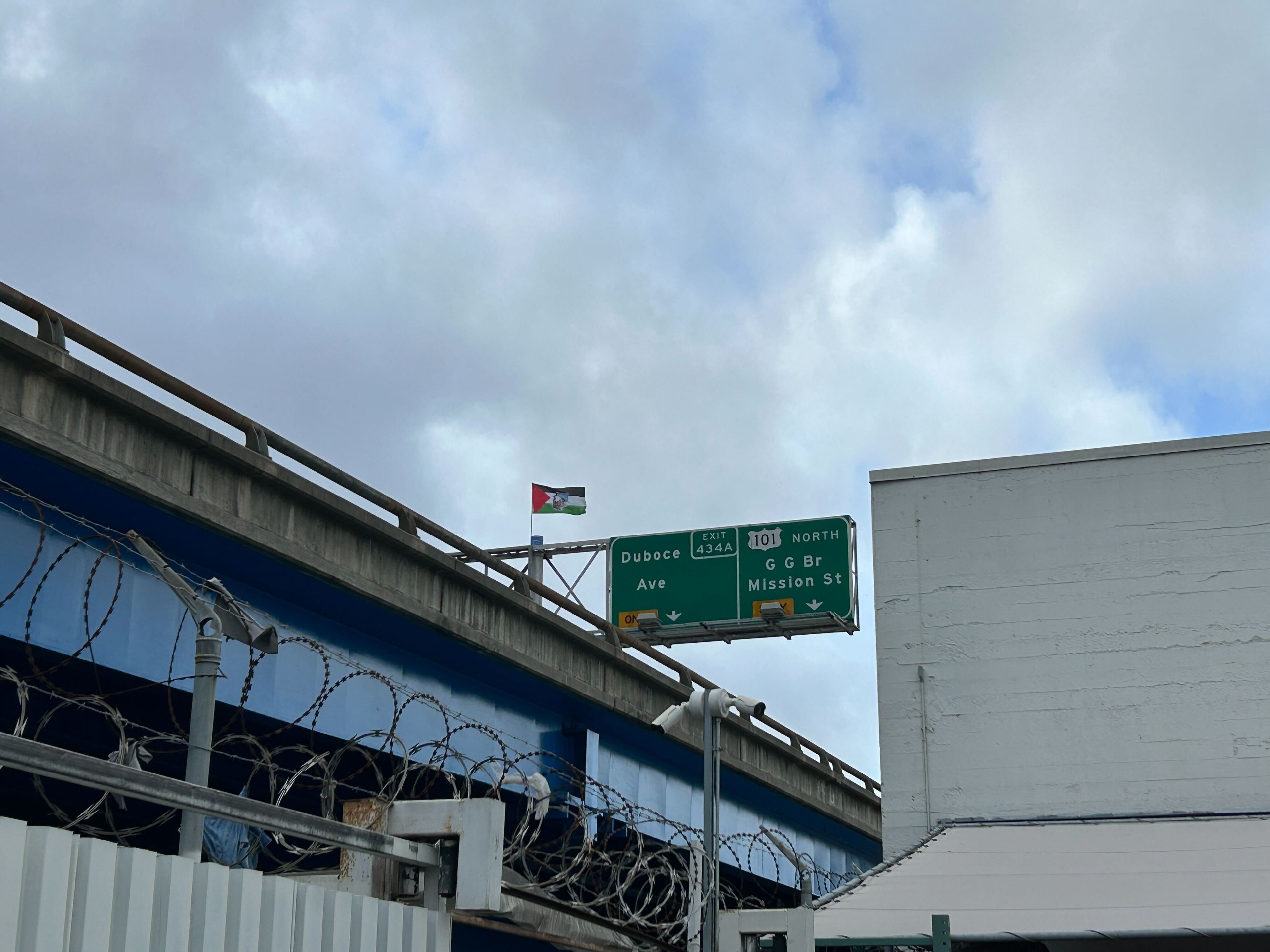 A flag stands above a railing on an elevated freeway under mostly cloudy skies
