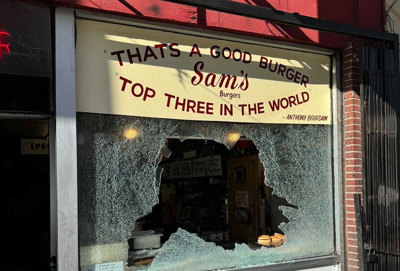 A shattered window with a sign above, reading "THAT'S A GOOD BURGER" and "TOP THREE IN THE WORLD - ANTHONY BOURDAIN" at "Sam's Burgers."