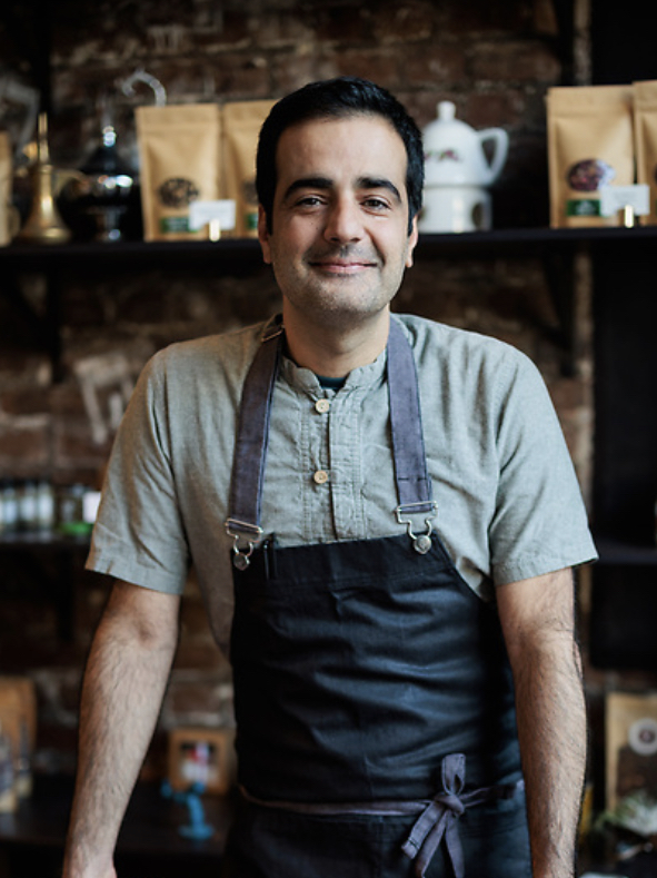 A smiling man in a grey shirt and dark apron stands in front of a coffee shelf.