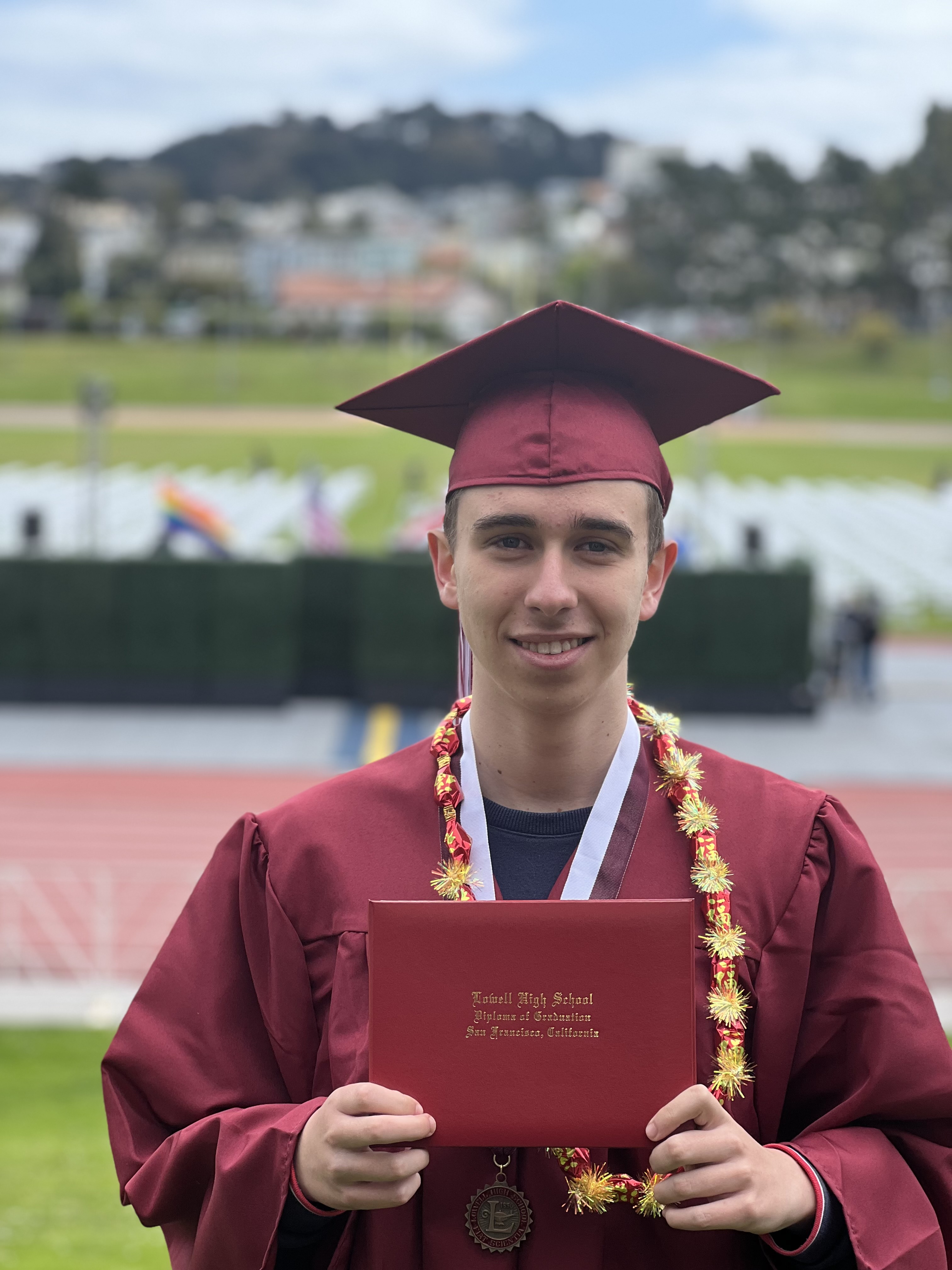 A young graduate in a maroon cap and gown holding a diploma, wearing a lei and medal, with a blurred background.
