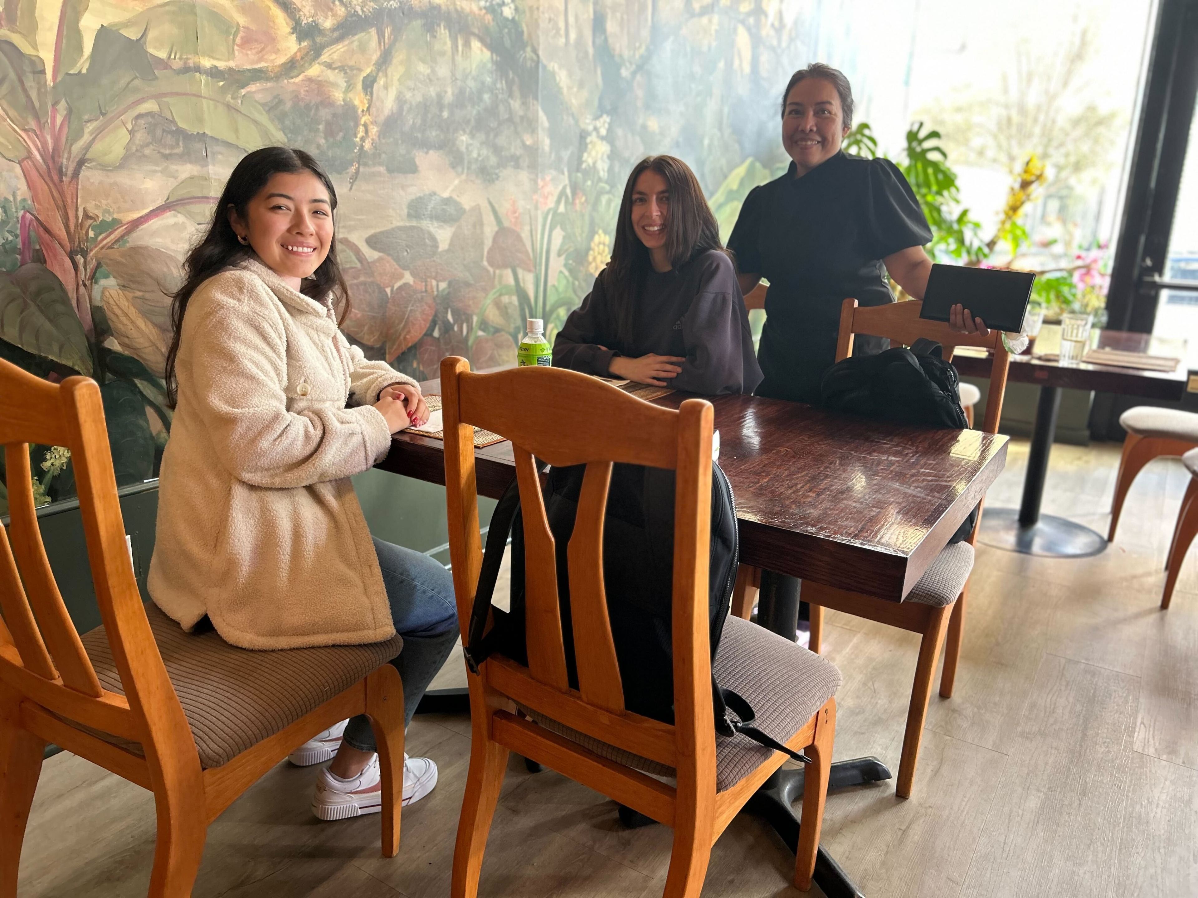 Three happy people are seated at a wooden table in a cozy café with a mural behind them.