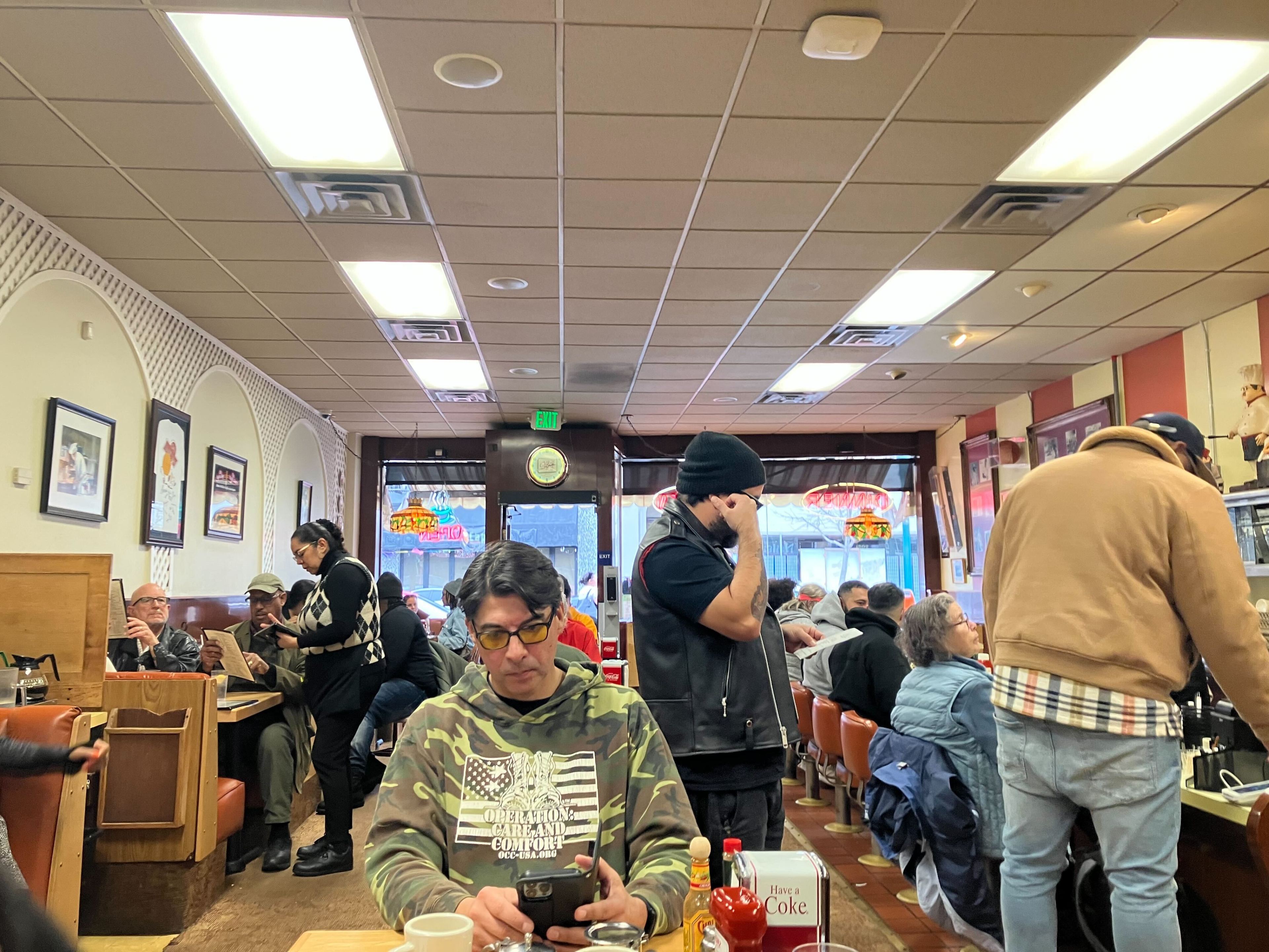 A bustling diner interior with patrons seated and dining, wall art, and a person checking their phone in the foreground.
