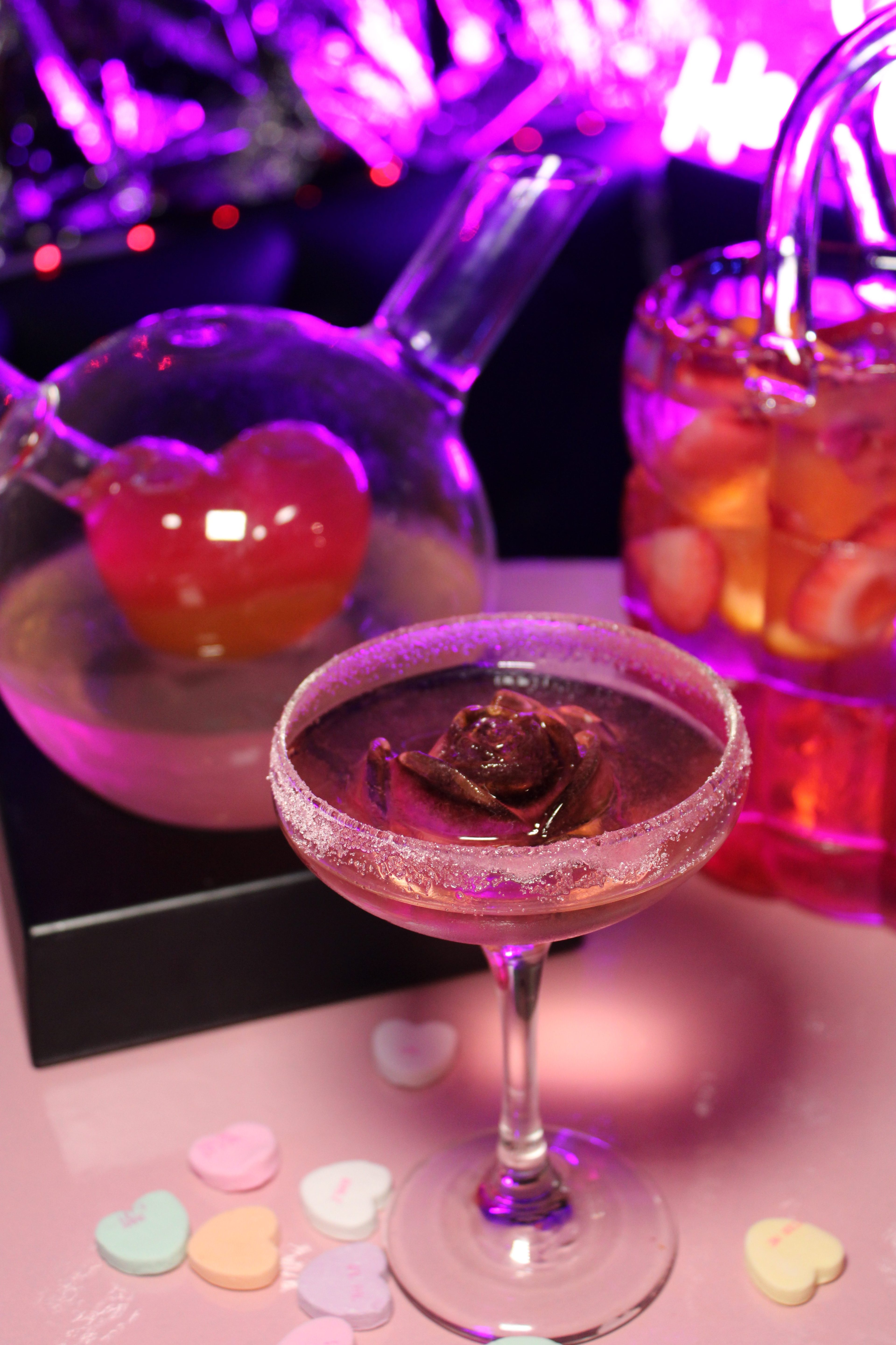 A red rose ice cub sits in a cocktail glass with candy hearts strewn below it.