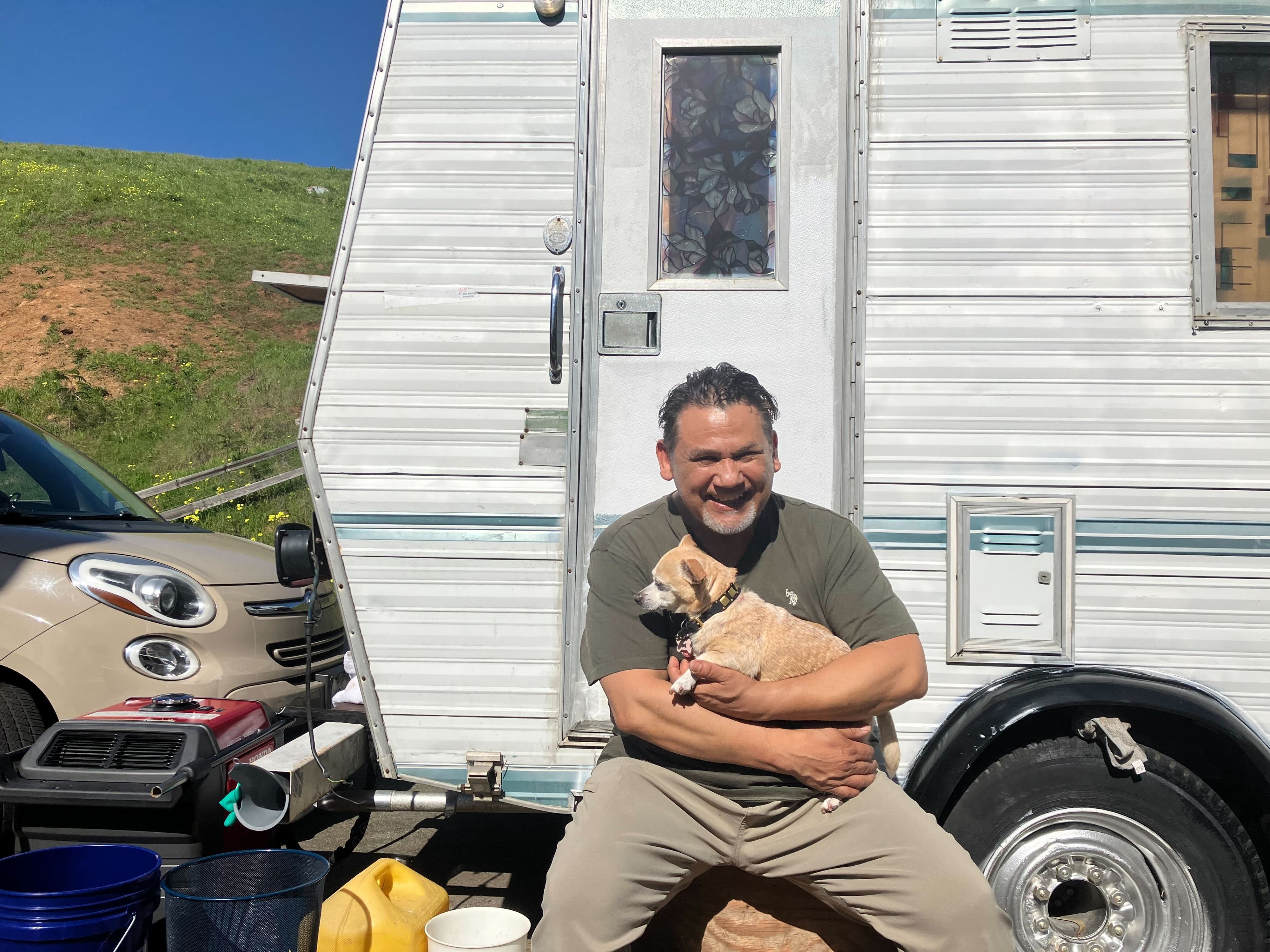 A smiling person sits beside an RV, holding a small dog, with a car and green hill in the background.