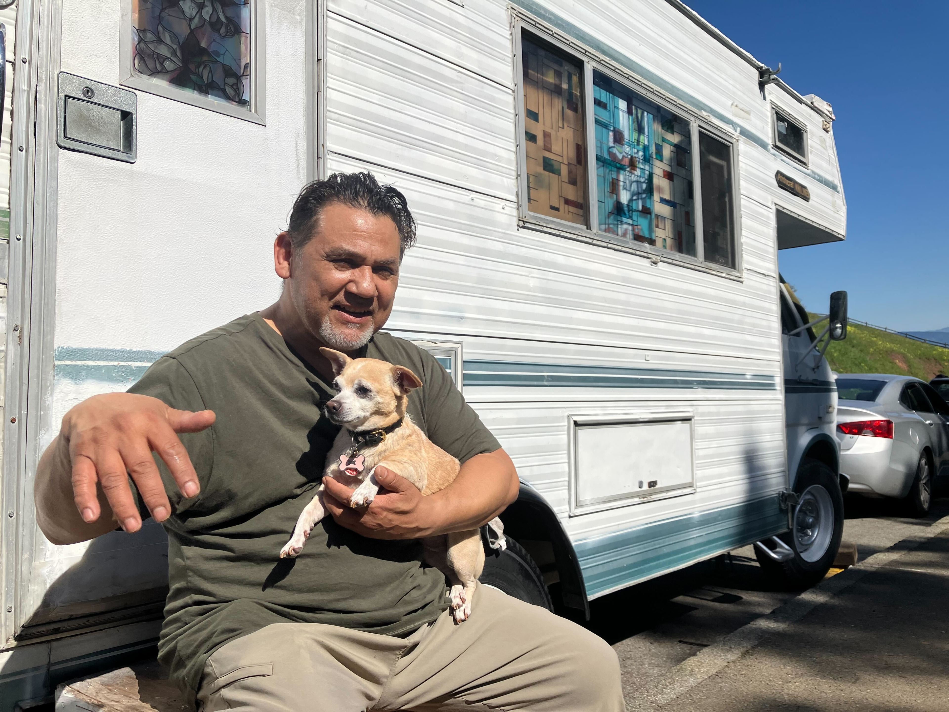 Man holds dog while sitting in front of white RV parked along street