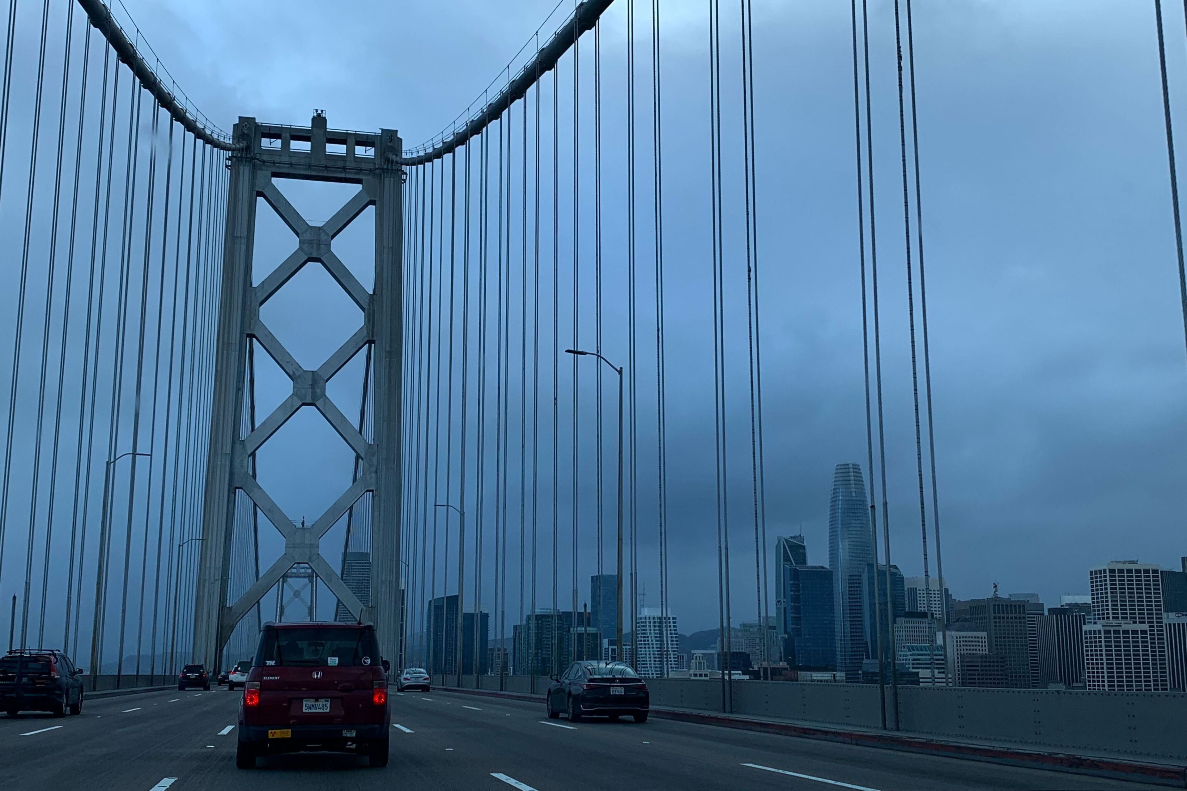 A city skyline is visible from a suspension bridge's traffic lanes under cloudy skies.