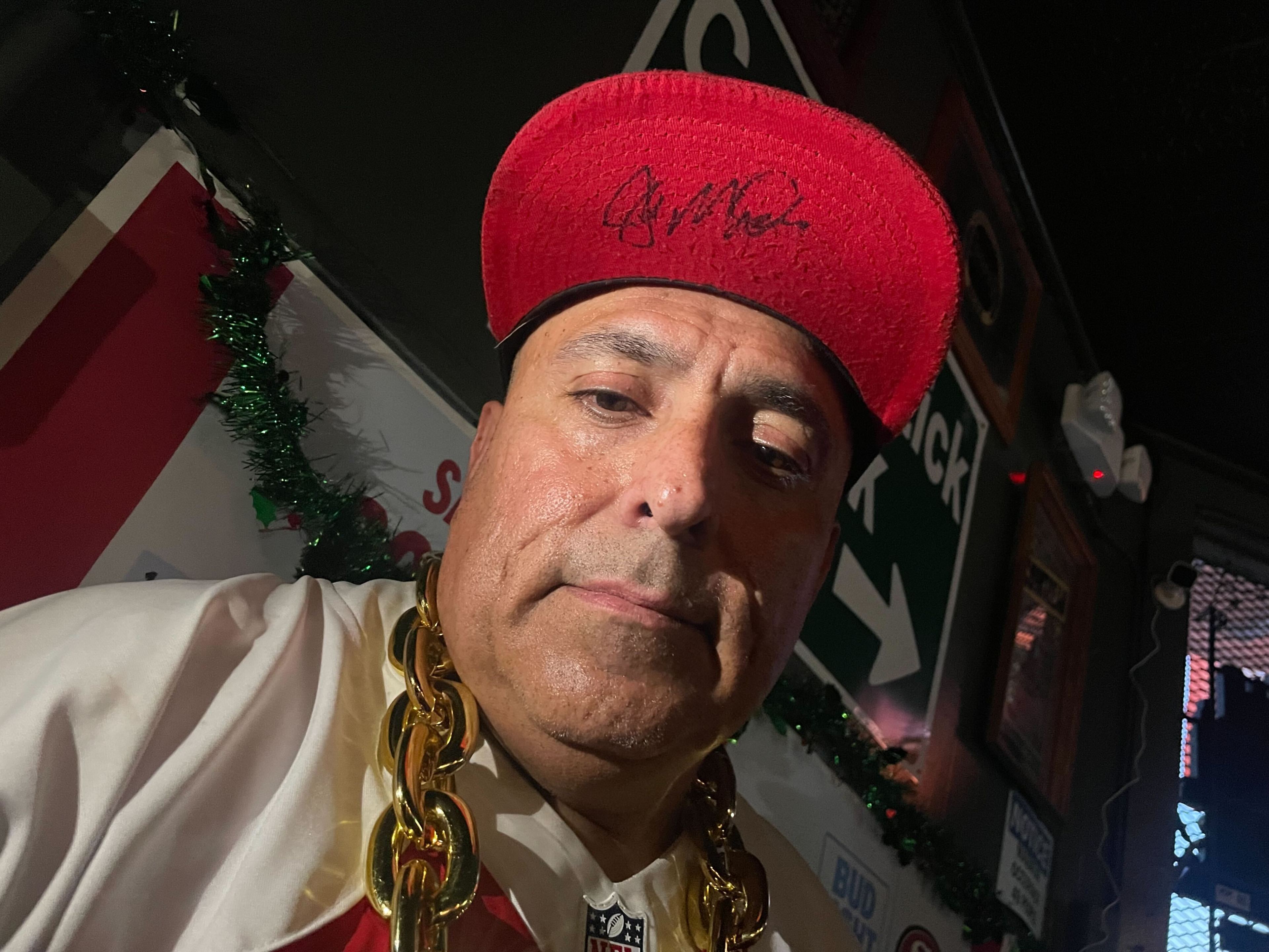 A man in a red cap and gold chain necklace is posing for a selfie with a bar interior background.