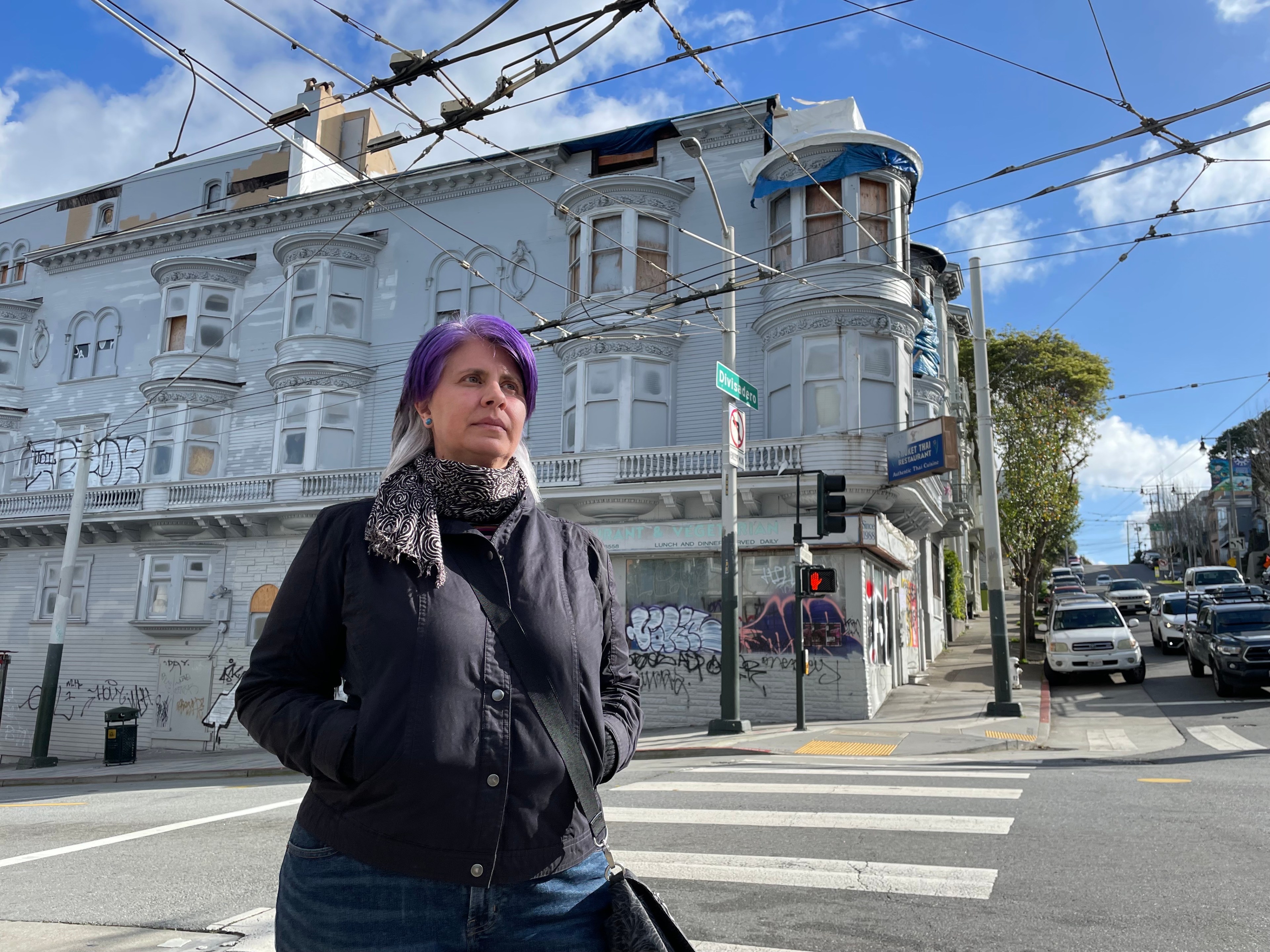 A woman stands on a street corner in front of a vacant, tagged-up four-story apartment building.
