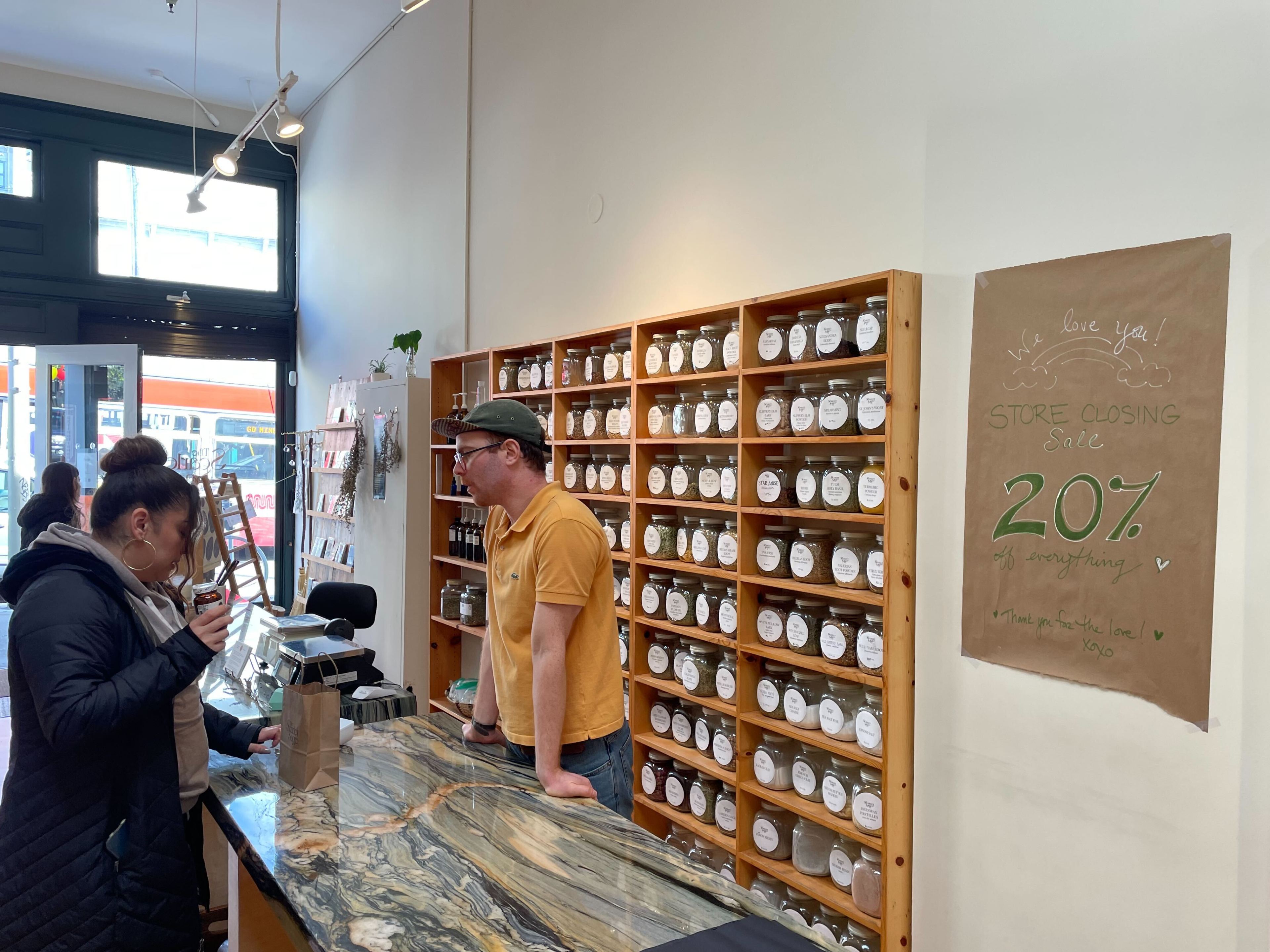 A shelf full of dried tea leaves is shown on sale for 20% off as a staff member and a customer stand nearby,