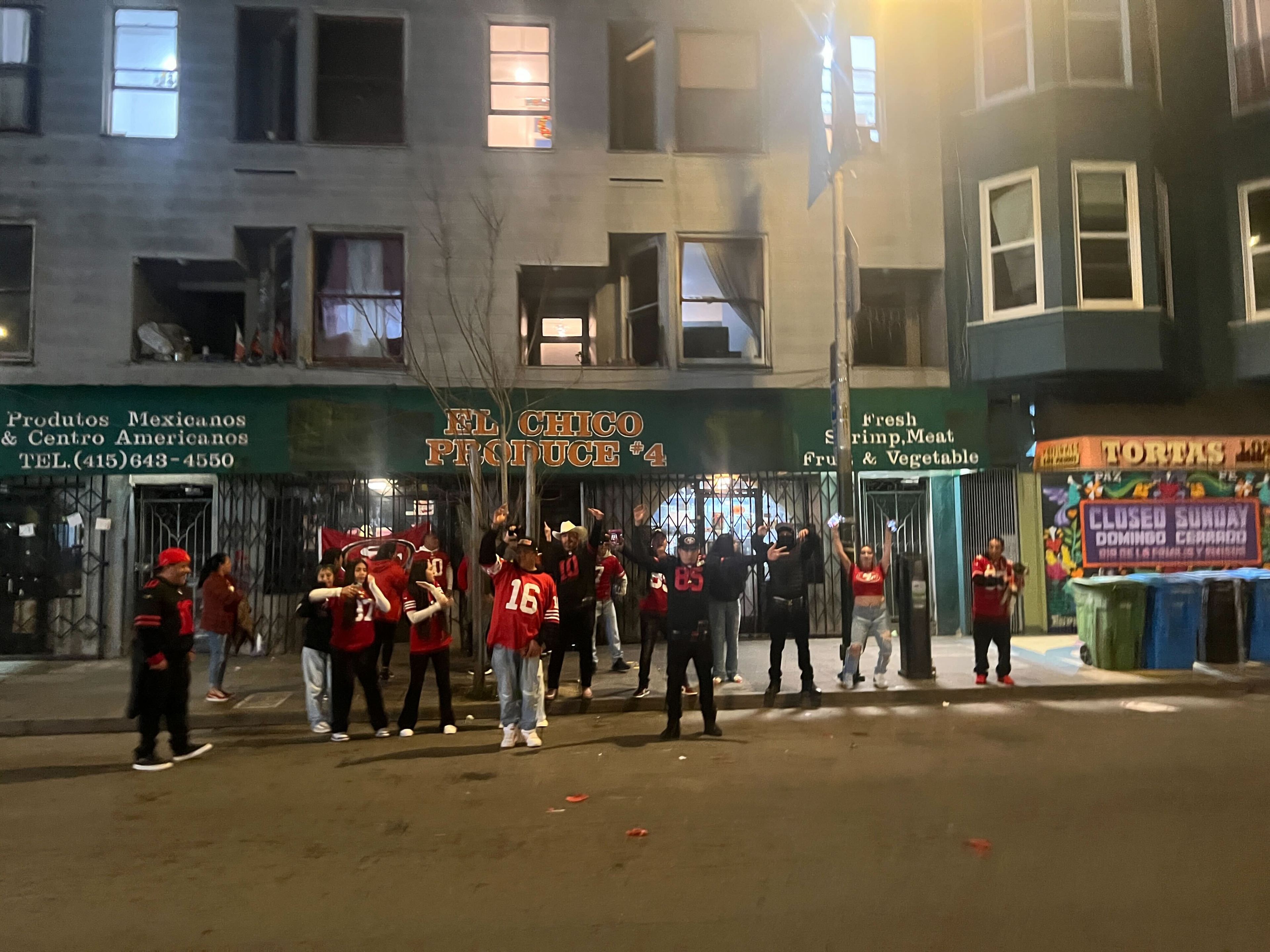 People in red jerseys are gathered outside shops on a city street at dusk. Signs indicate Latin American products.