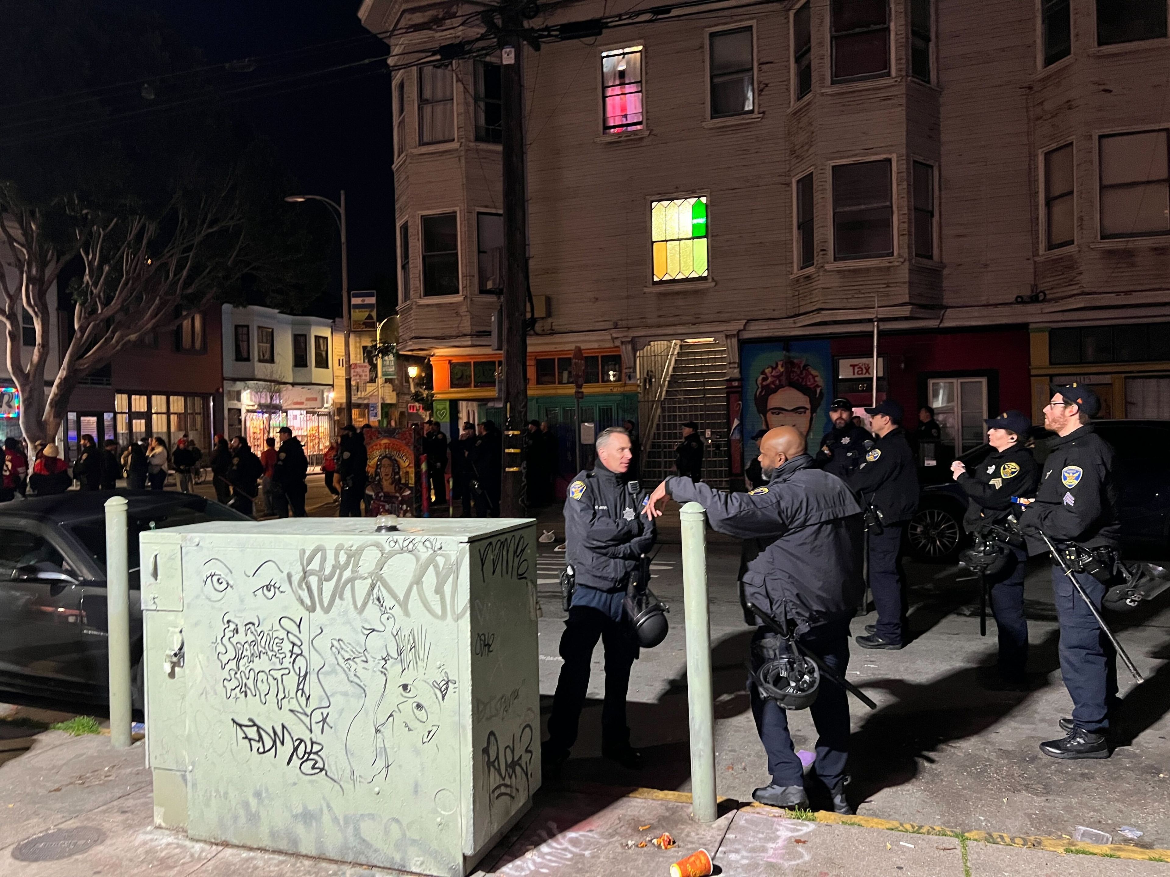 Police officers are standing by a graffiti-covered utility box at night, with onlookers and city buildings in the background.