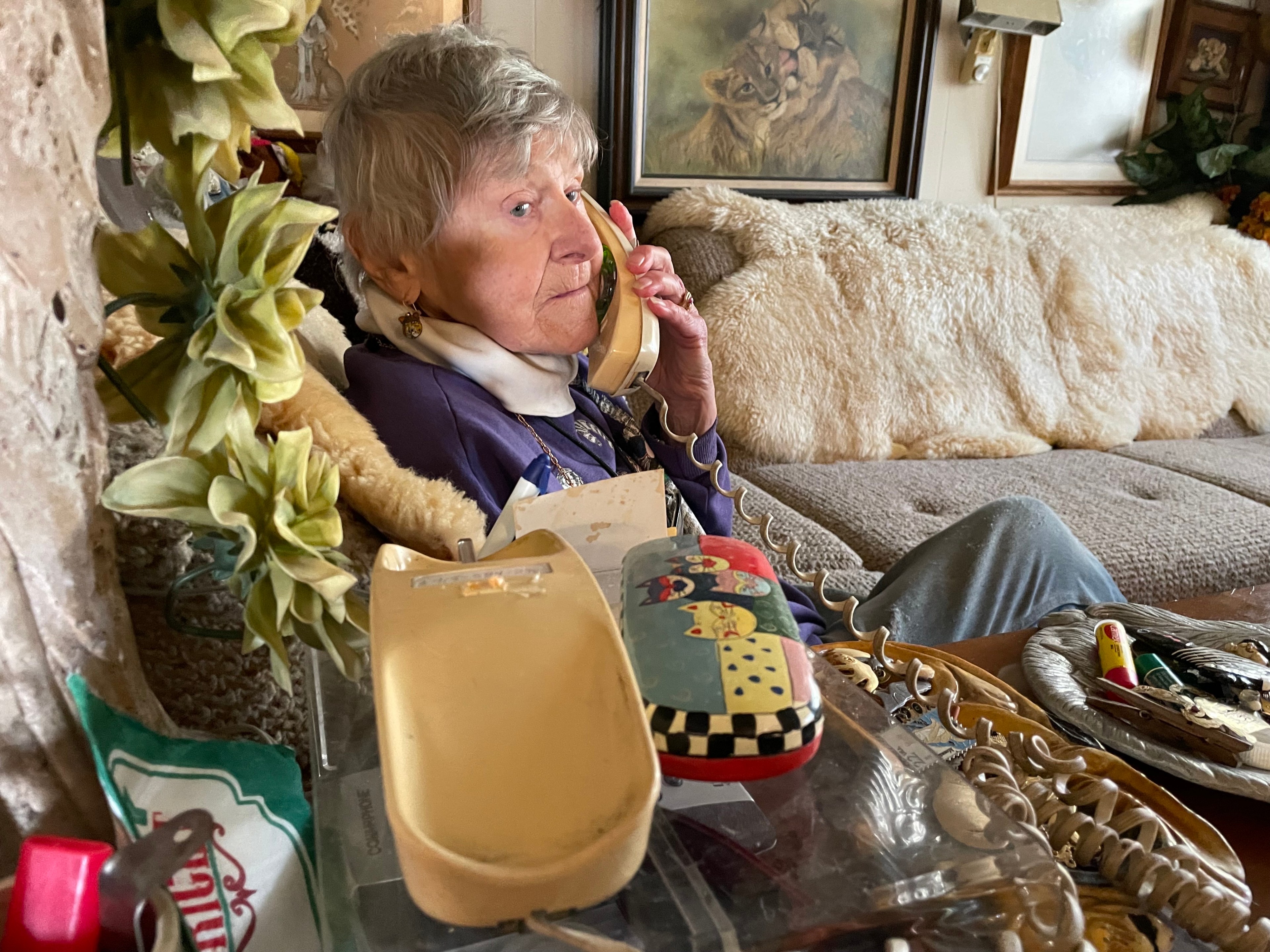 An old woman sits on a section sofa holding a corded landline telephone.