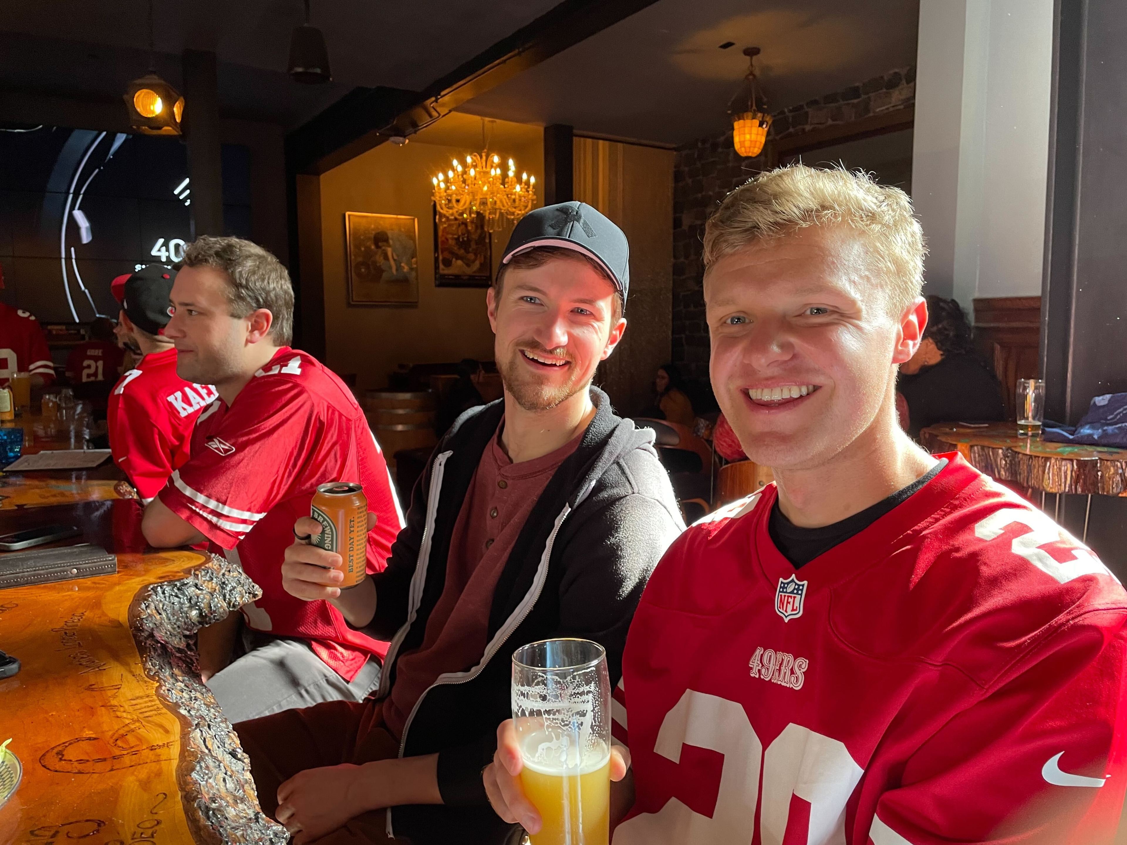 Three men in a pub, two smiling at the camera, one with a beer, in a cheerful atmosphere with sports on TV.