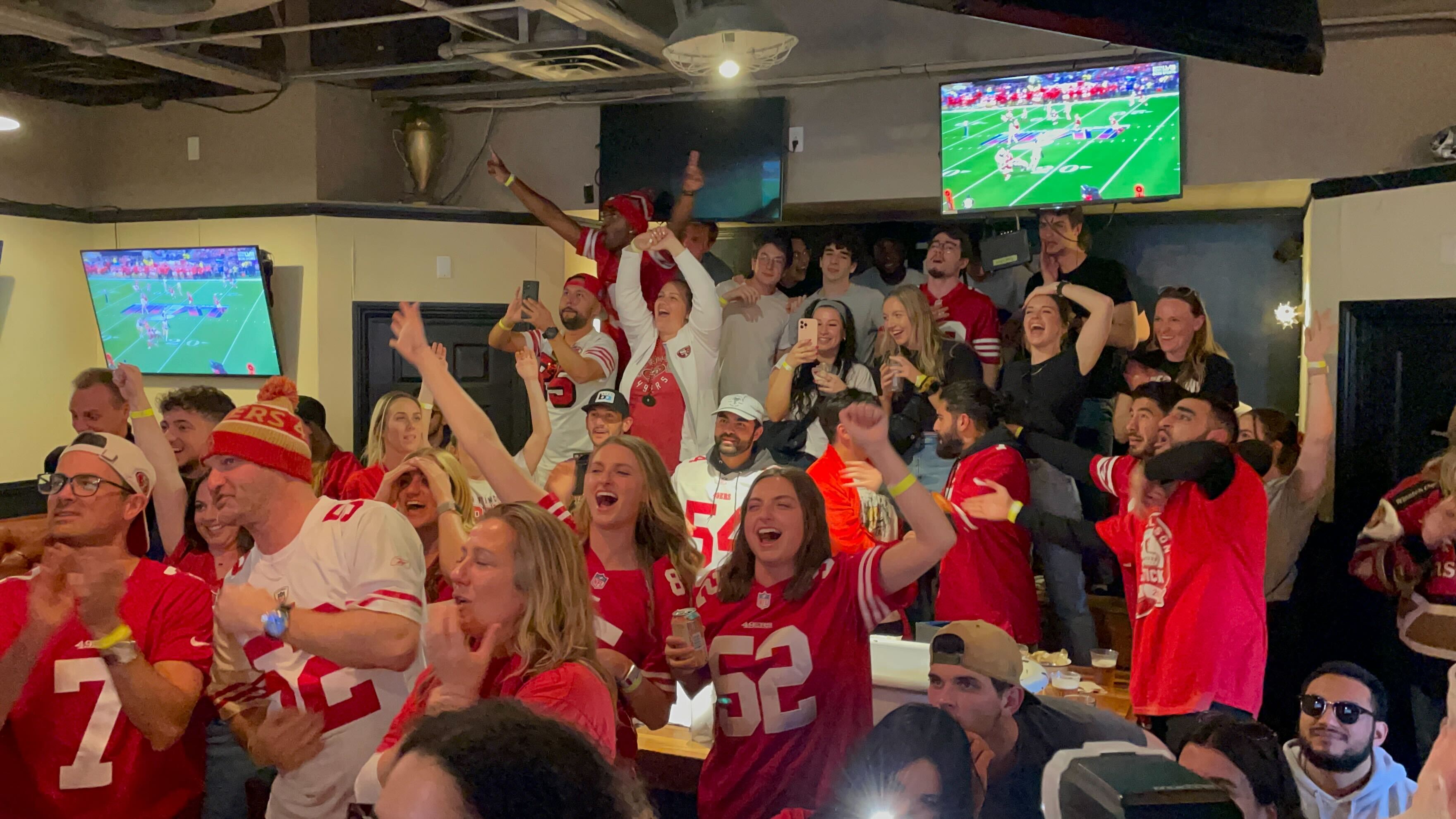A lively crowd in sports jerseys cheering in a bar with TVs showing a football game.