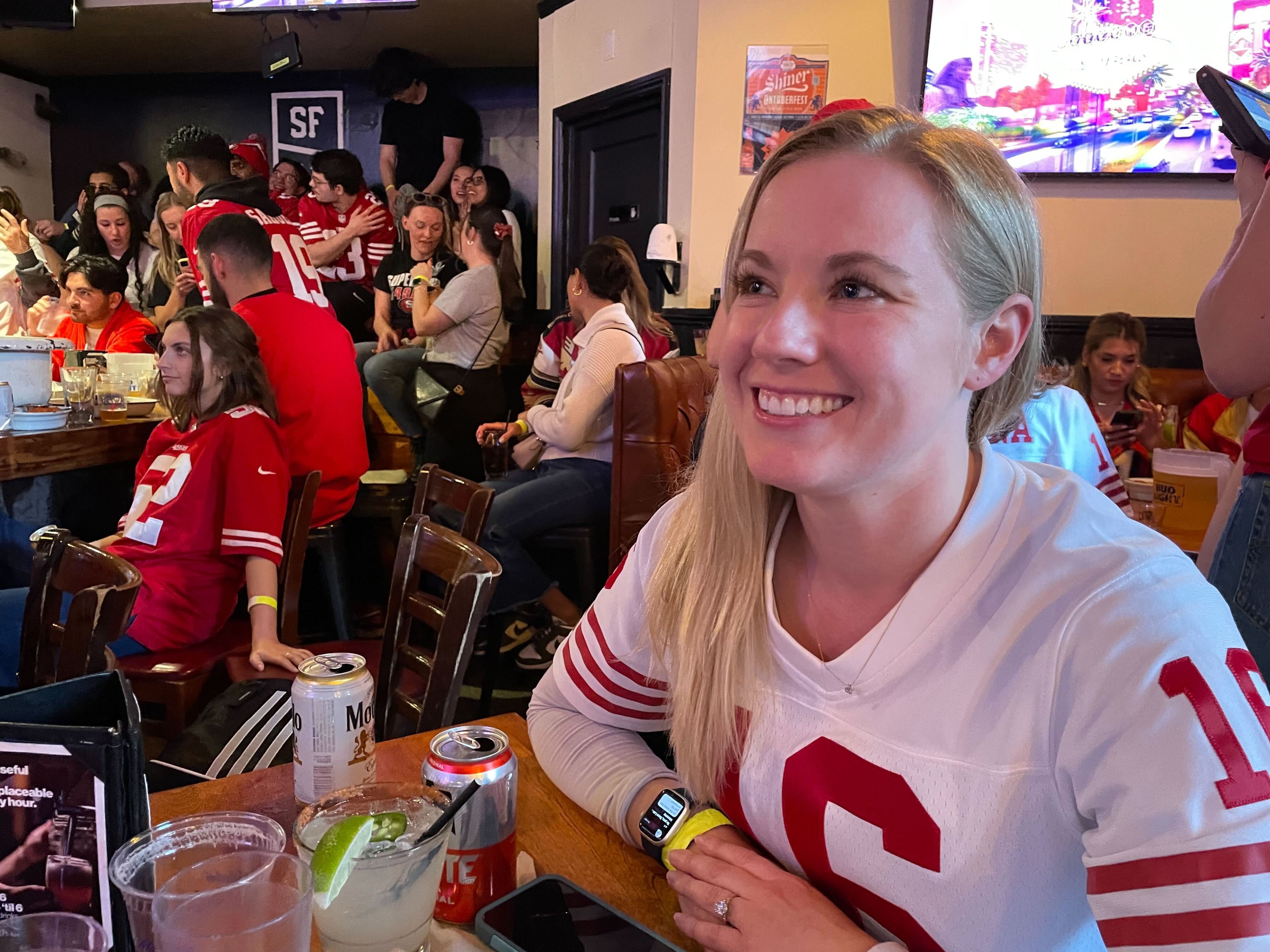 A smiling woman in a number 10 sports jersey sits in a bar filled with people wearing red and white.