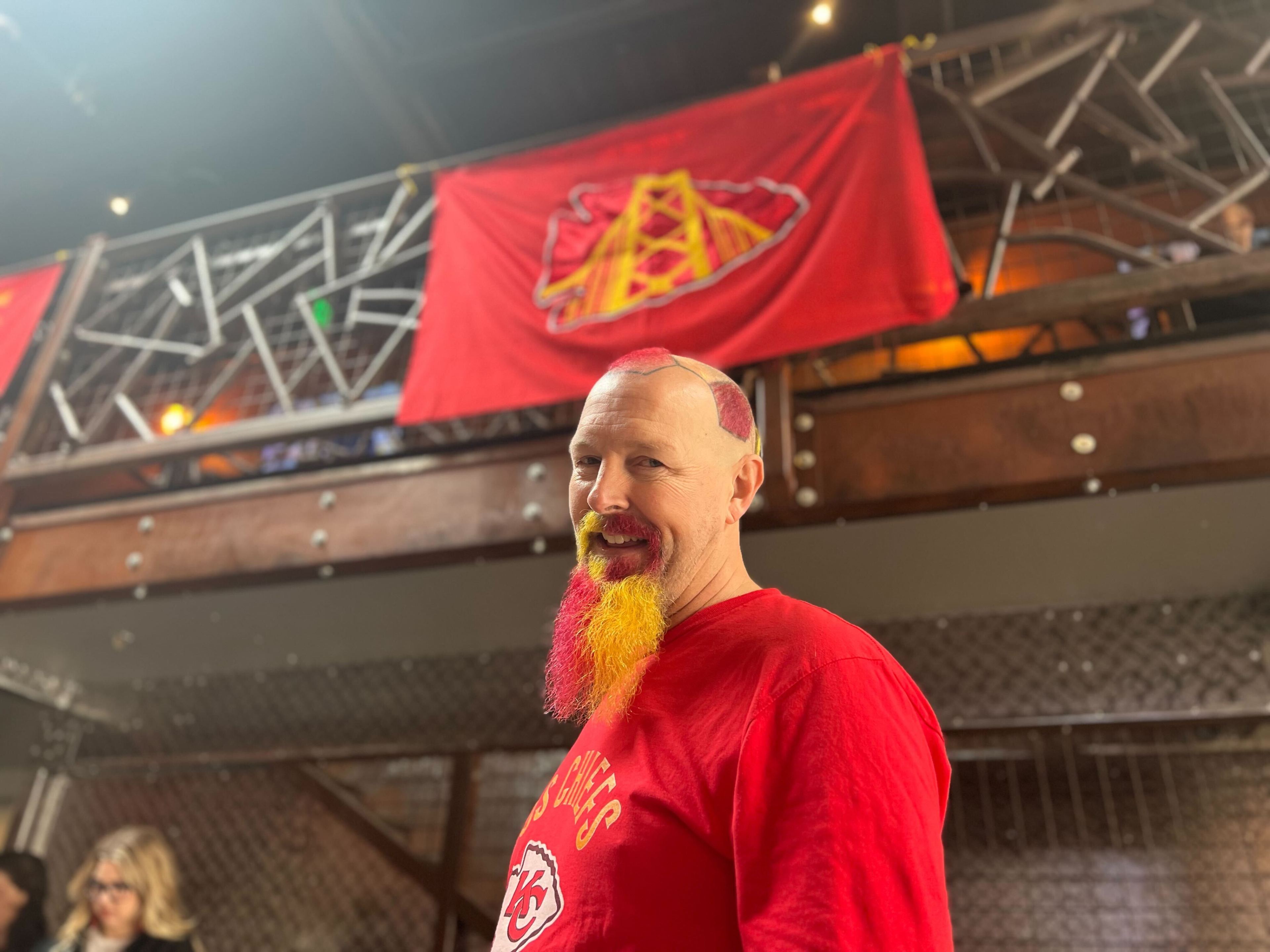 A man with a dyed red and yellow beard smiles, wearing a red shirt with &quot;Chiefs&quot; on it; a team flag hangs in the background.