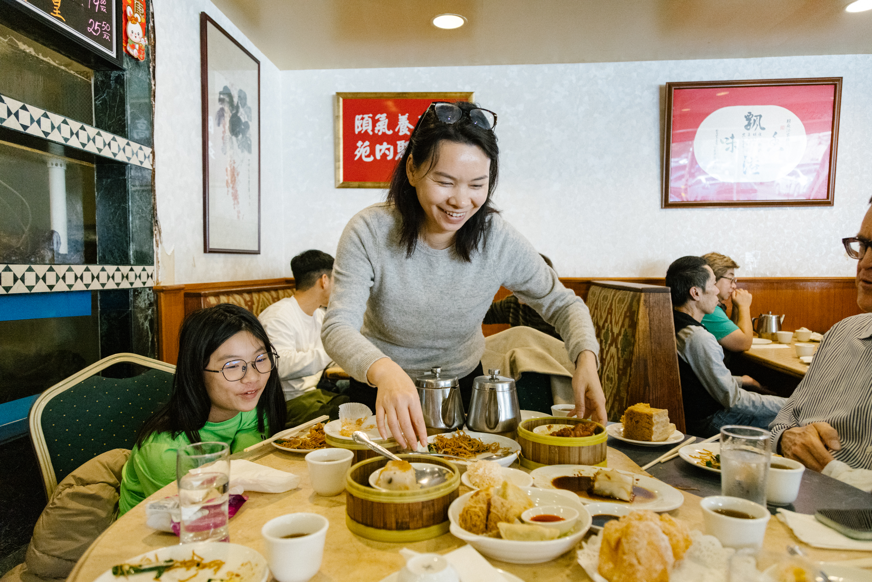 A woman serves dim sum at a busy restaurant table where a child looks on eagerly. Cups of tea and various Chinese dishes abound.