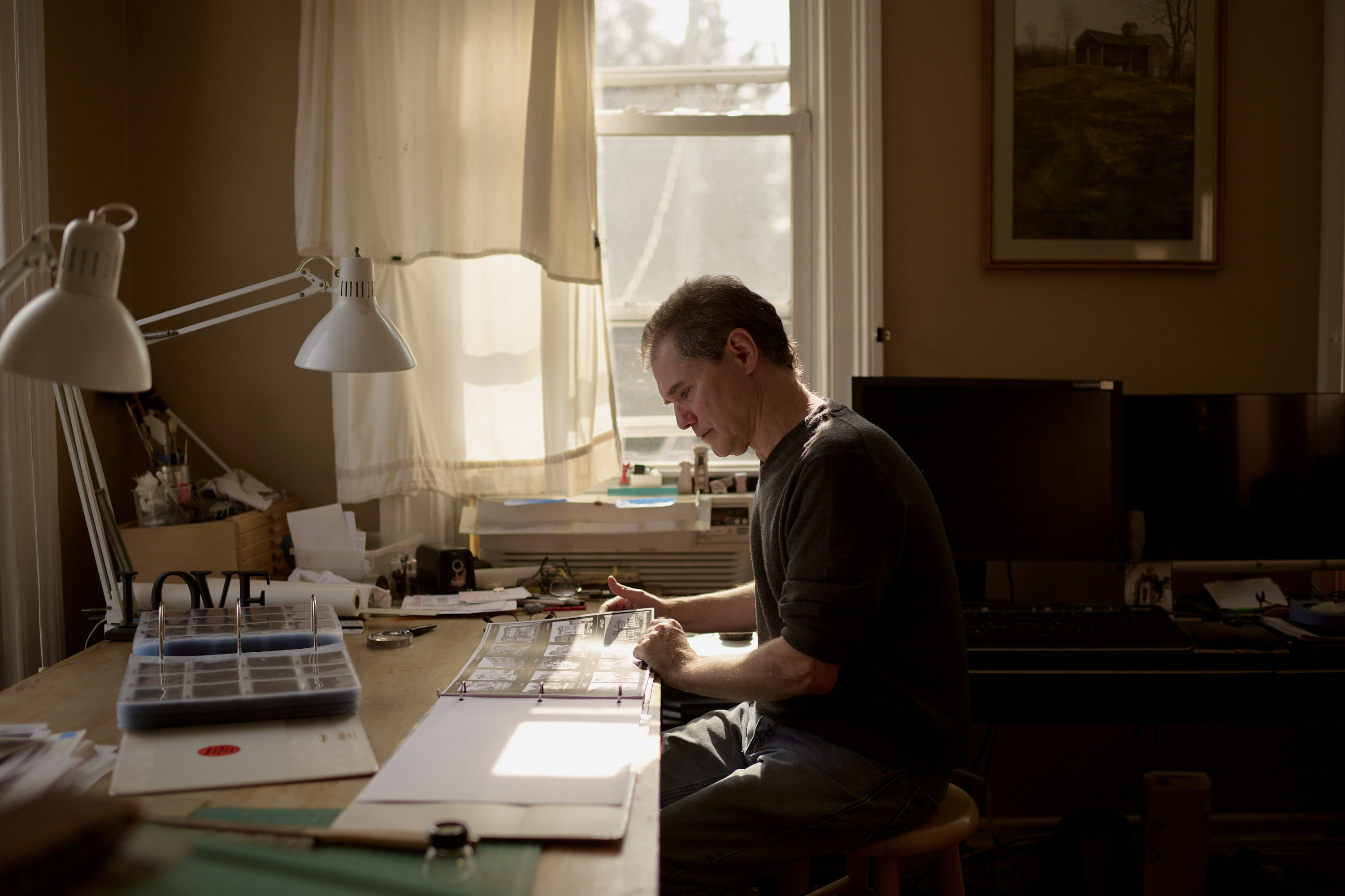 A person is focused on reviewing documents at a cluttered desk with warm lighting from a window.