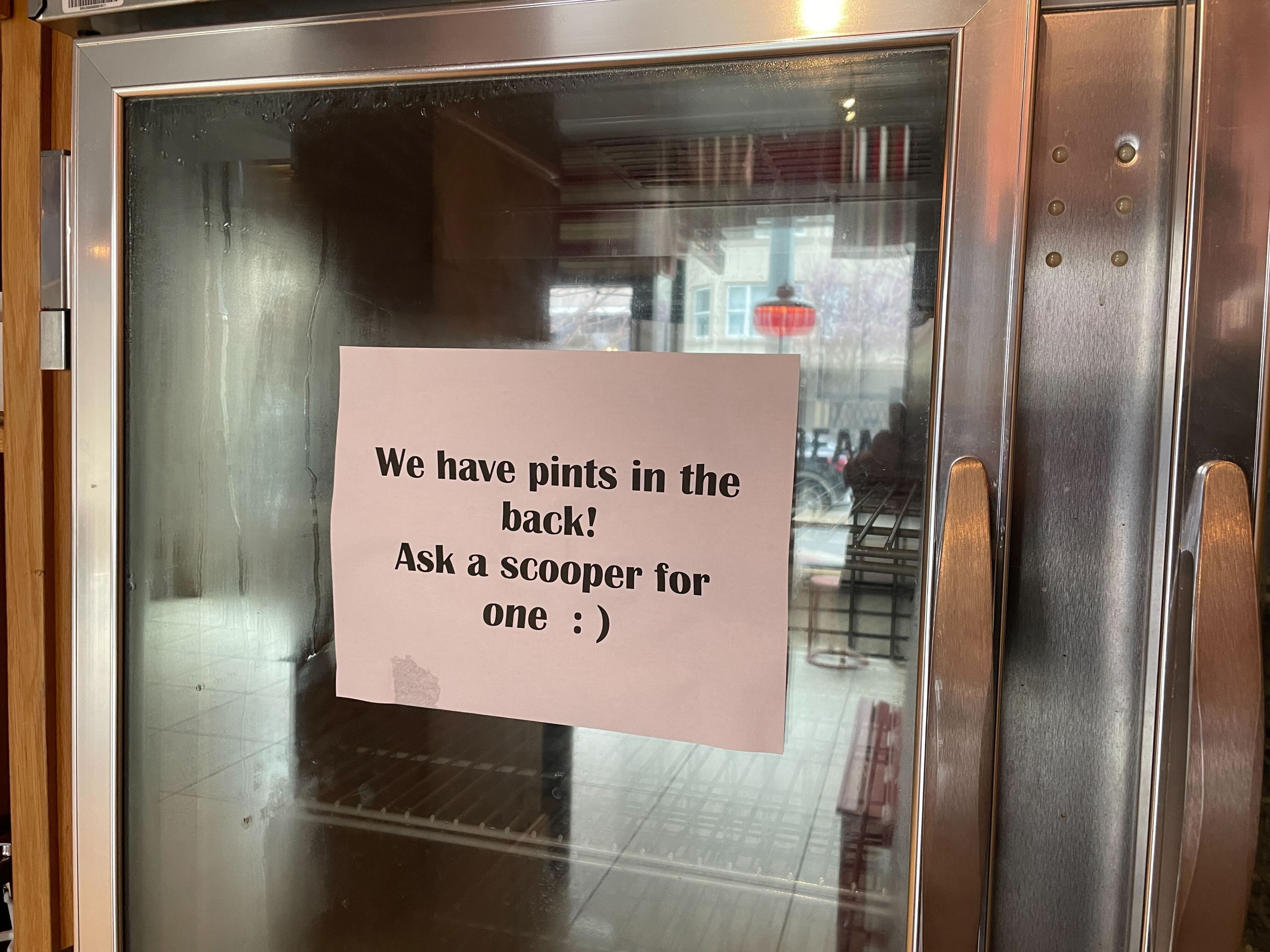 A sign on a freezer door reads &quot;We have pints in the back! Ask a scooper for one :)&quot; reflecting a casual, inviting tone.