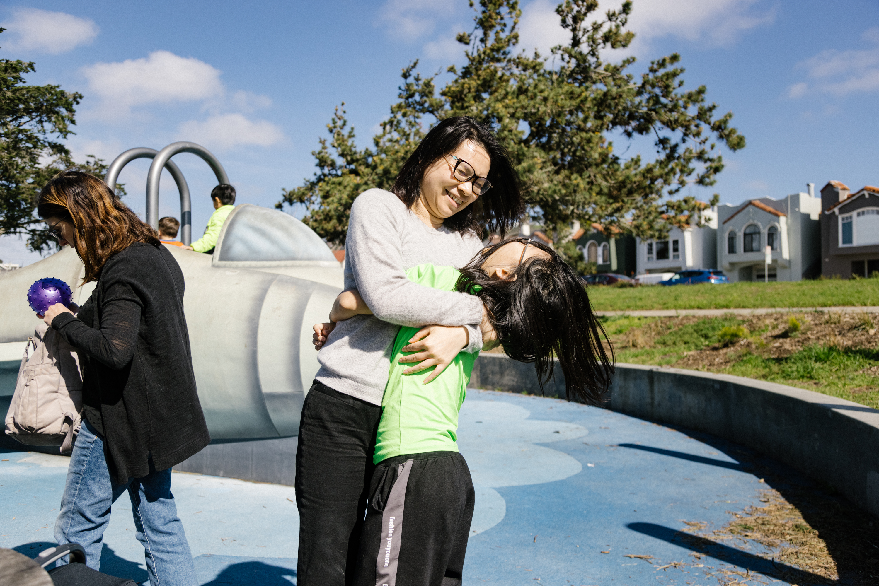 A woman hugs a joyful child at a sunny park, with others and houses in the background.