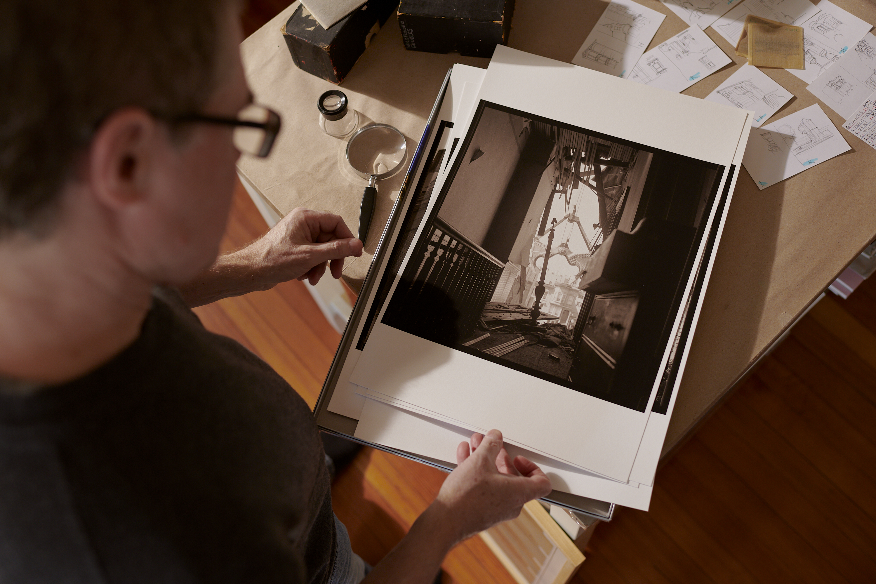 A person examines a black-and-white photograph of a staircase amidst debris; sketches and a magnifying glass lay nearby.