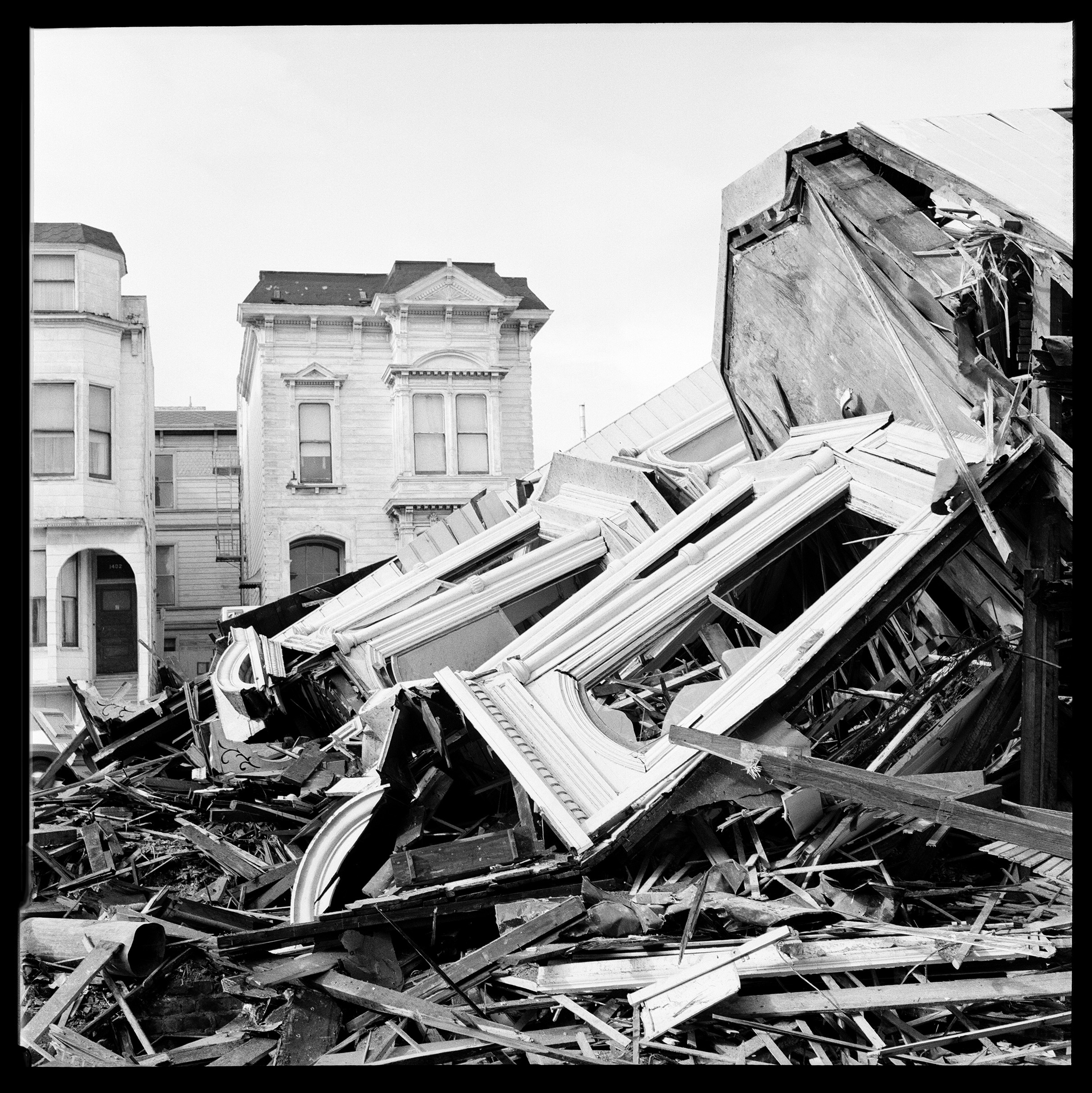 A black and white photo of destroyed wooden structures in a heap, with intact Victorian buildings in the background.
