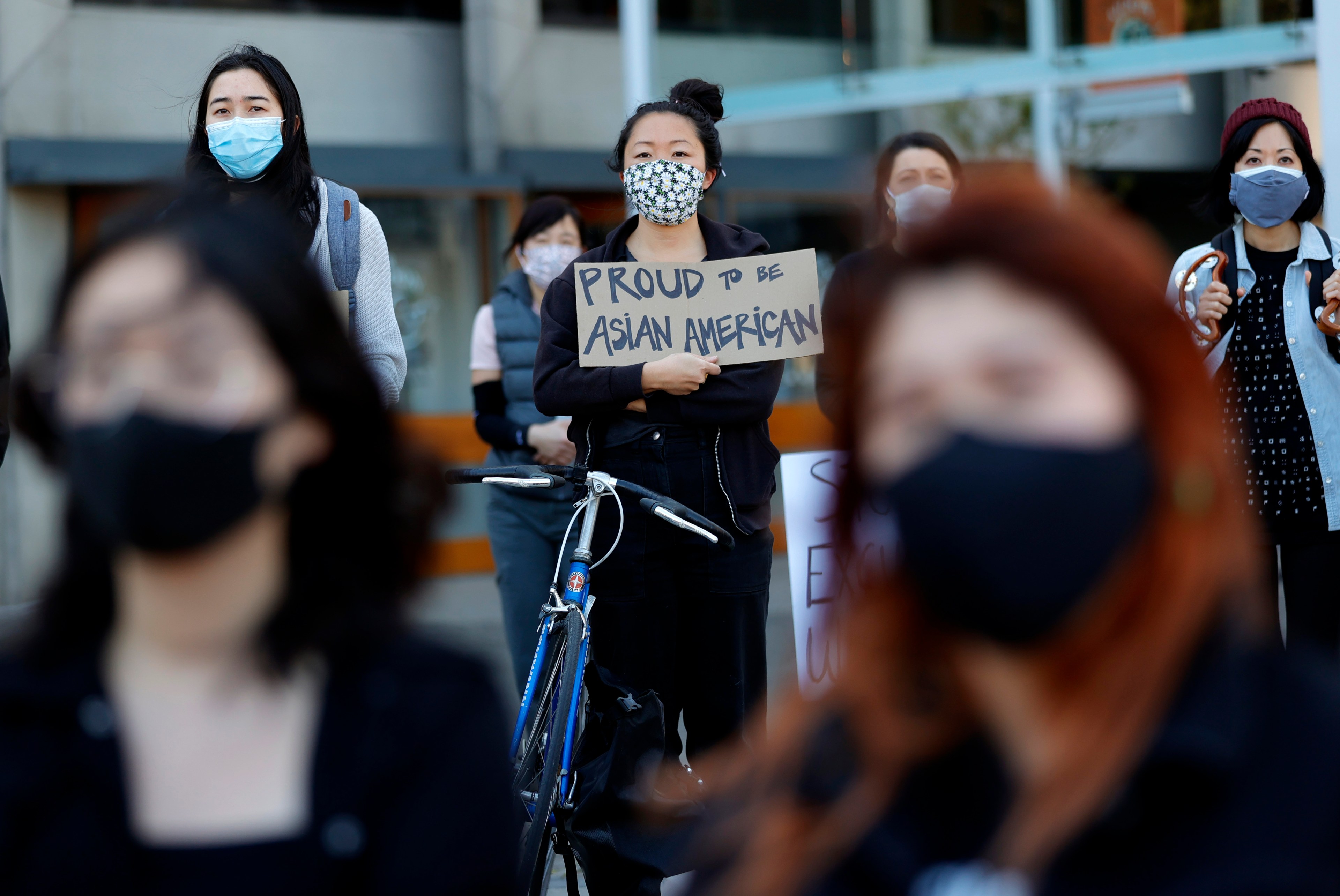 People in masks, one holding a &quot;PROUD TO BE ASIAN AMERICAN&quot; sign, seemingly at a gathering or protest.