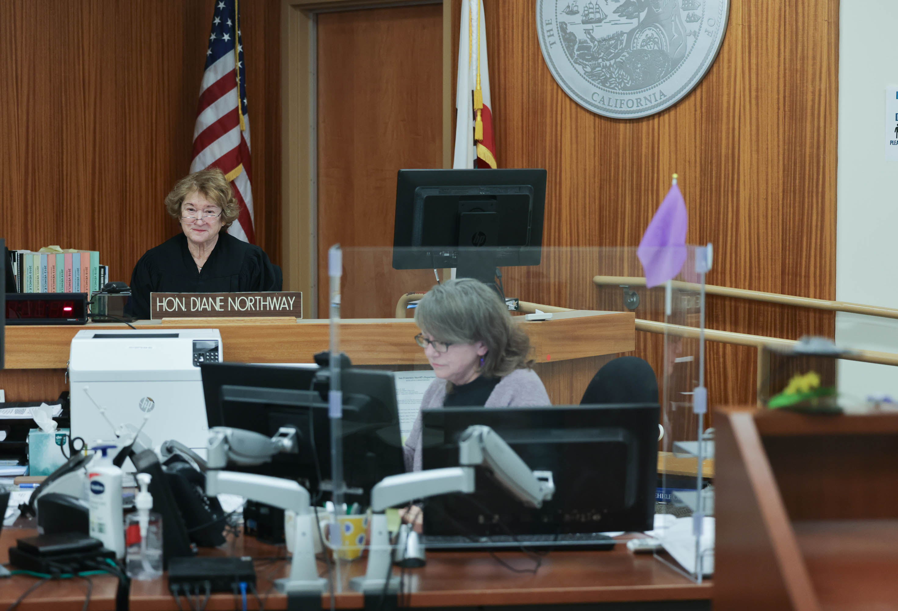 A judge sits on a dais in the background and a clerk sits at a computer in the foreground.
