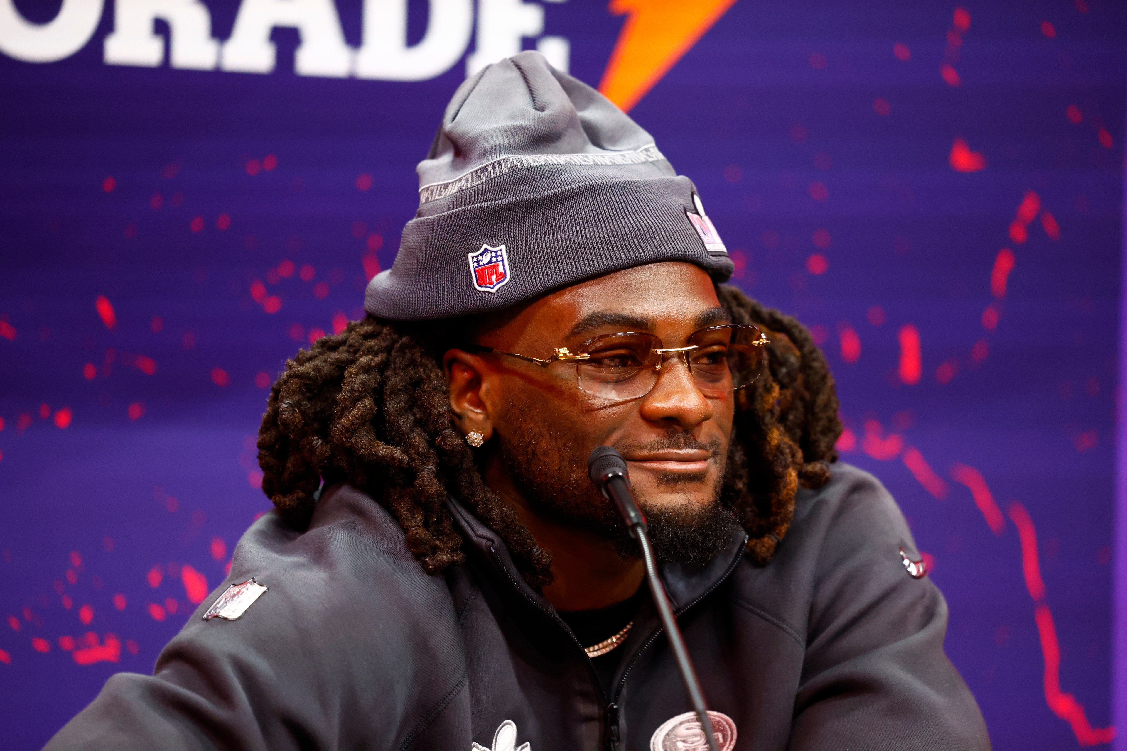 A person with dreadlocks and glasses sits smiling, wearing a beanie and sports jacket with an NFL logo against a purple backdrop.