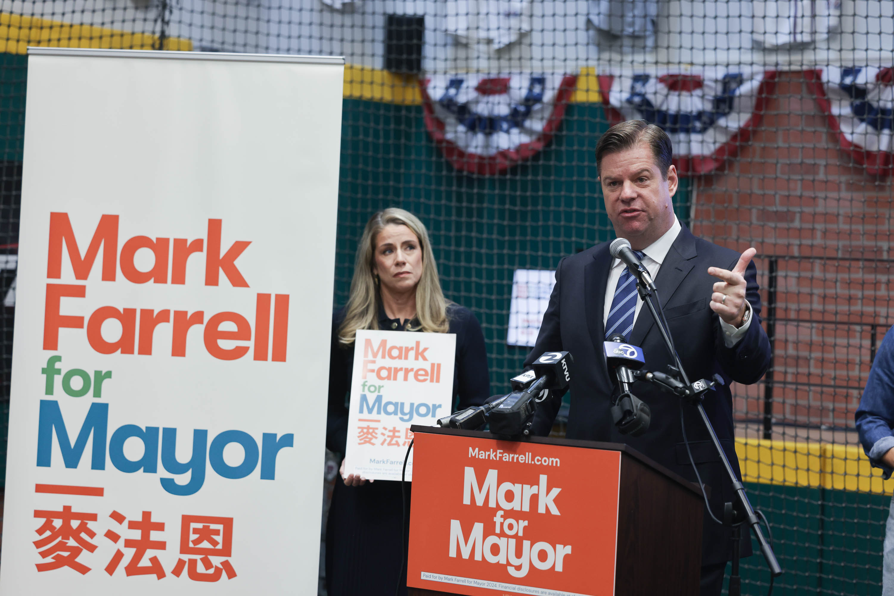 A man speaks at a podium with &quot;Mark Farrell for Mayor&quot; signs, a woman stands behind him, and microphones are in front.