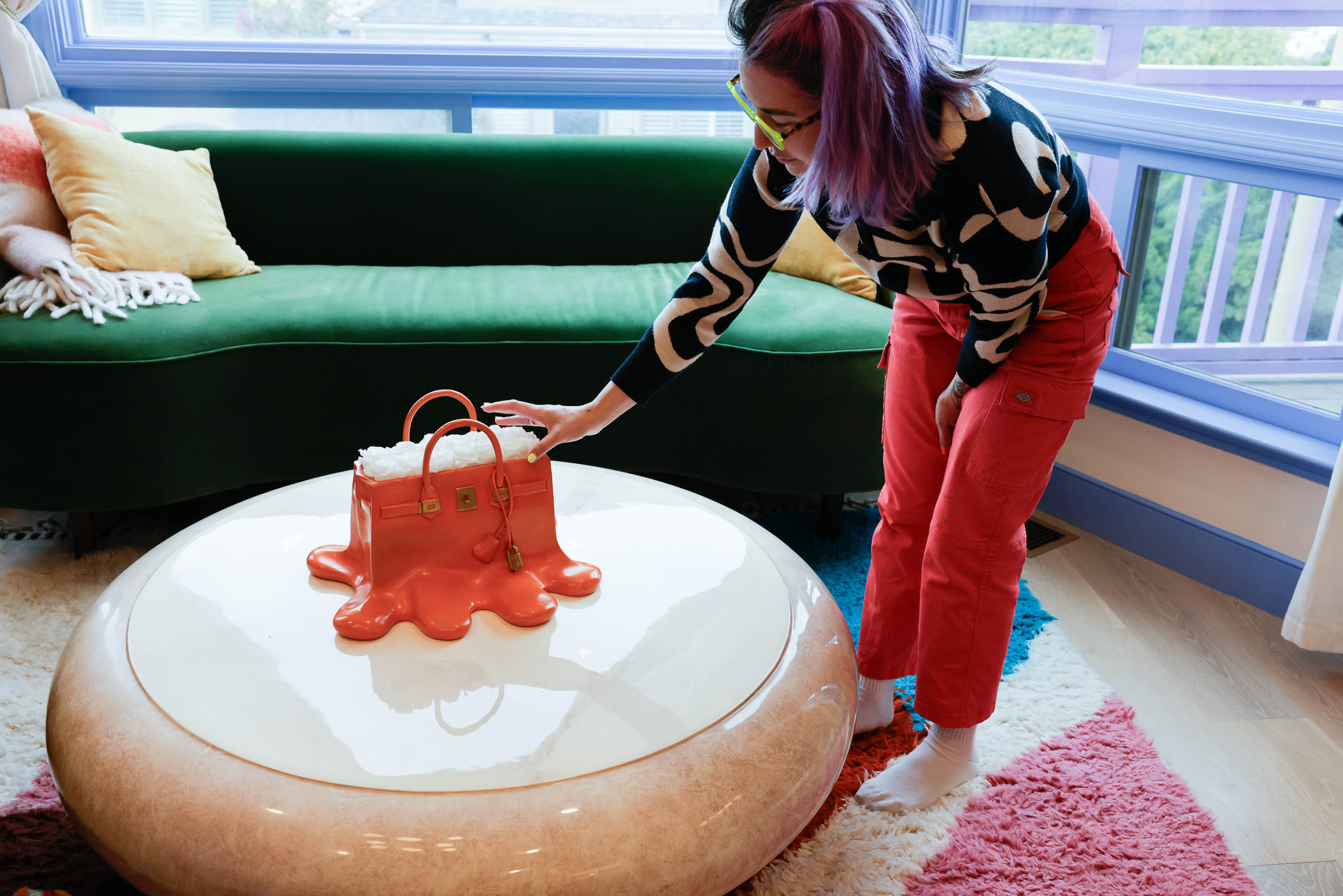A person with colorful hair touches a melting, red purse sculpture on a round table, beside a green sofa with pillows.