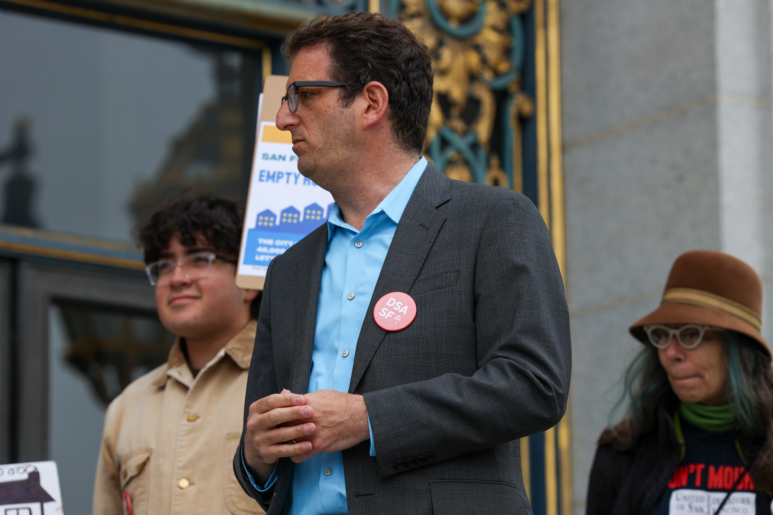 Dean Preston stands in profile with his hands clasped. He is wearing a blue dress shit and gray jacket and standing in front of a younger Latinx person holding a protest sign.
