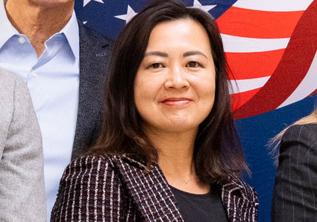 A woman smiling at the camera, with a US flag backdrop, wearing a plaid blazer. Partial views of others are seen.