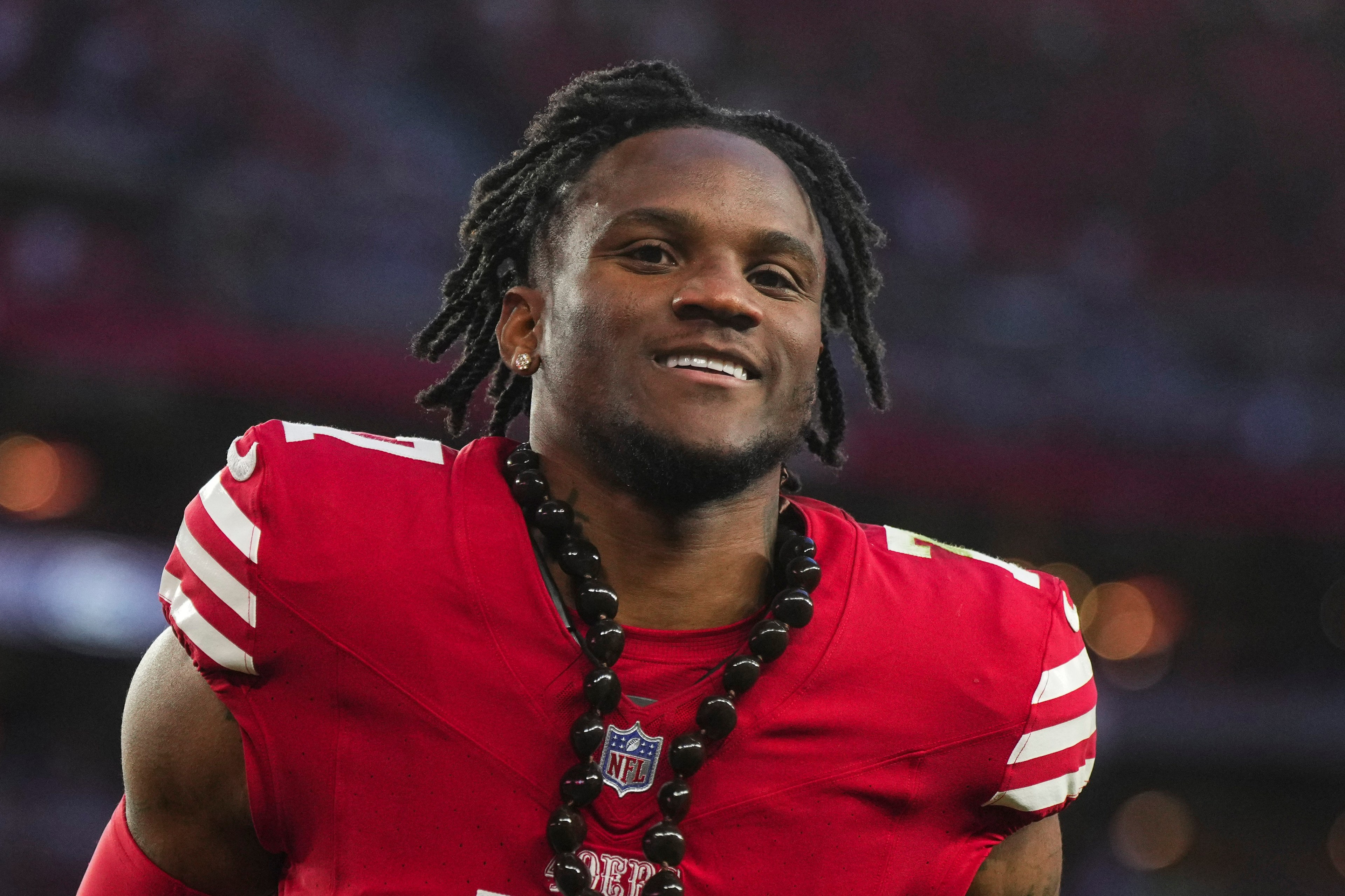 A smiling football player with dreadlocks in a red jersey adorned with a large bead necklace.