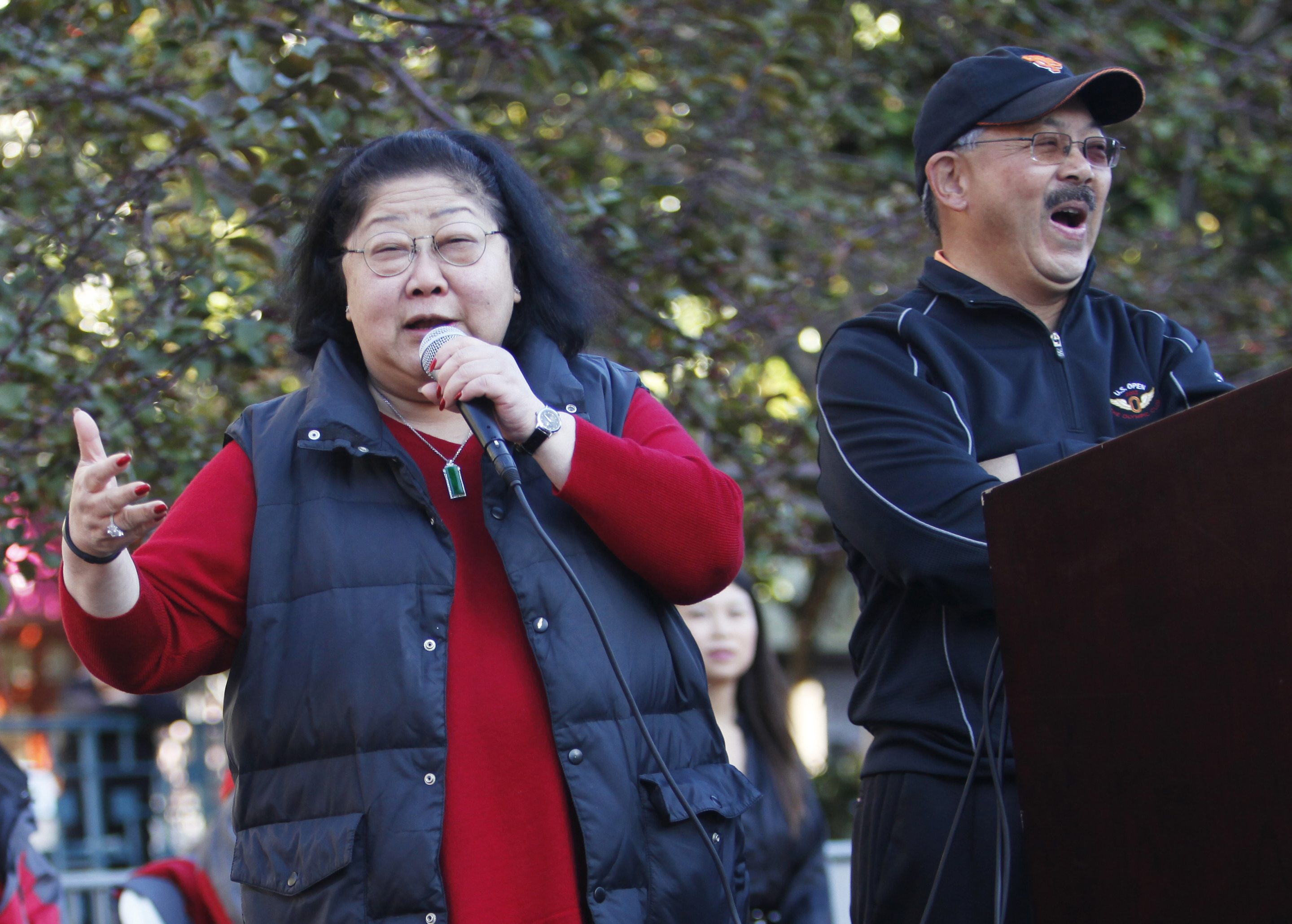 A woman speaks into a microphone, a smiling man beside her, with trees and a person in the background.