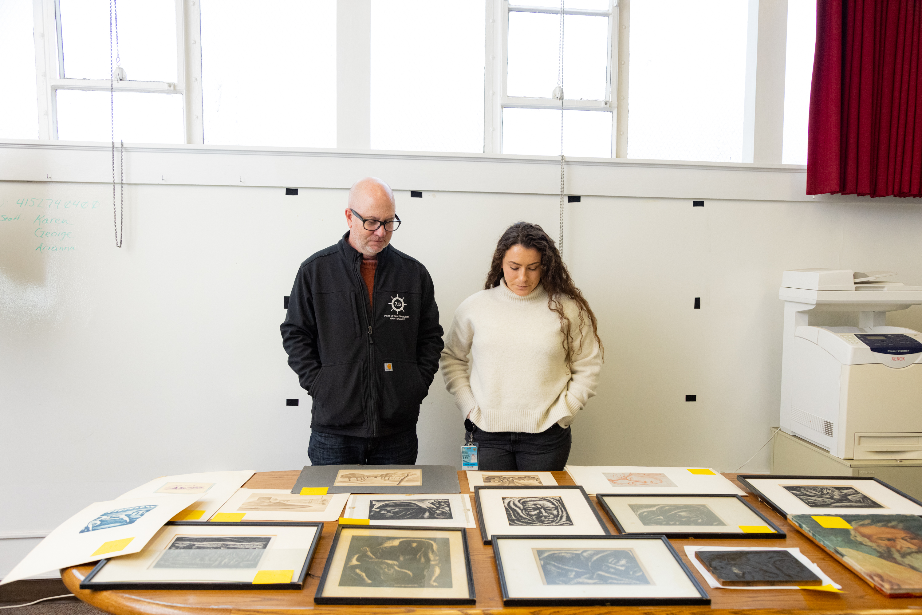 Two people are observing various framed artworks displayed on a table in a bright room.