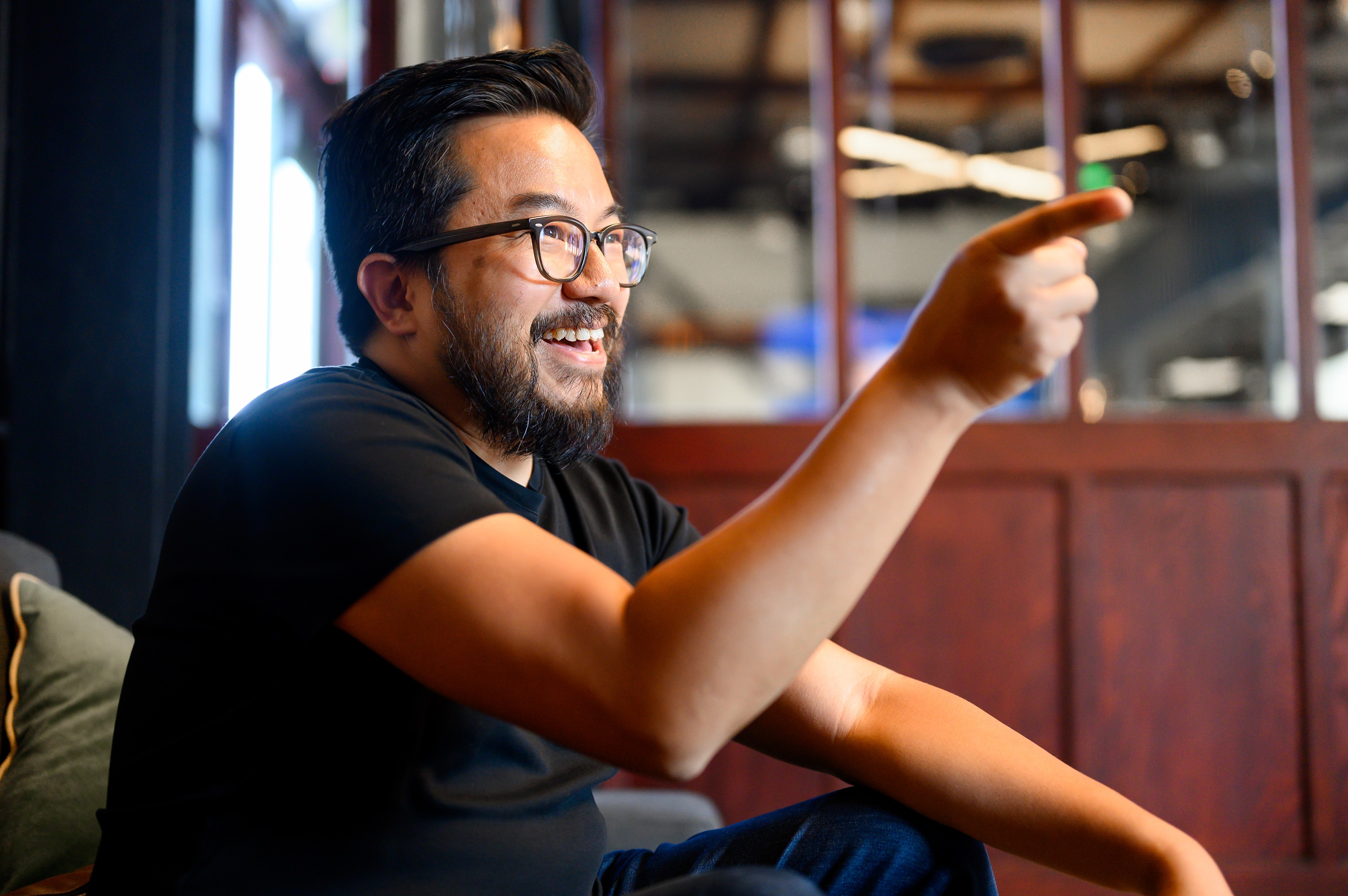 an asian man in a short-sleeved black shirt and dark-framed glasses faces right and points an index finger out of the frame.