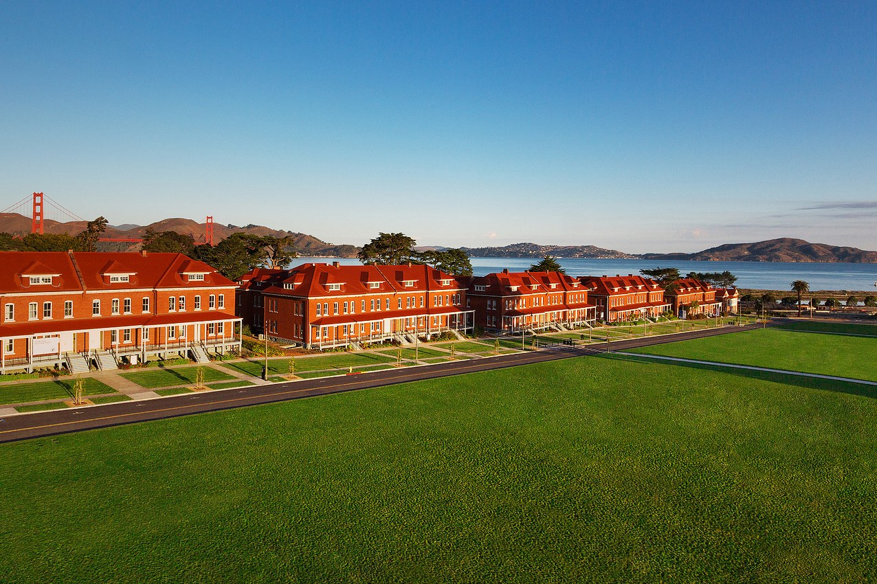Row of red-roofed buildings with Golden Gate Bridge in the distance, beside a green lawn under clear blue skies.
