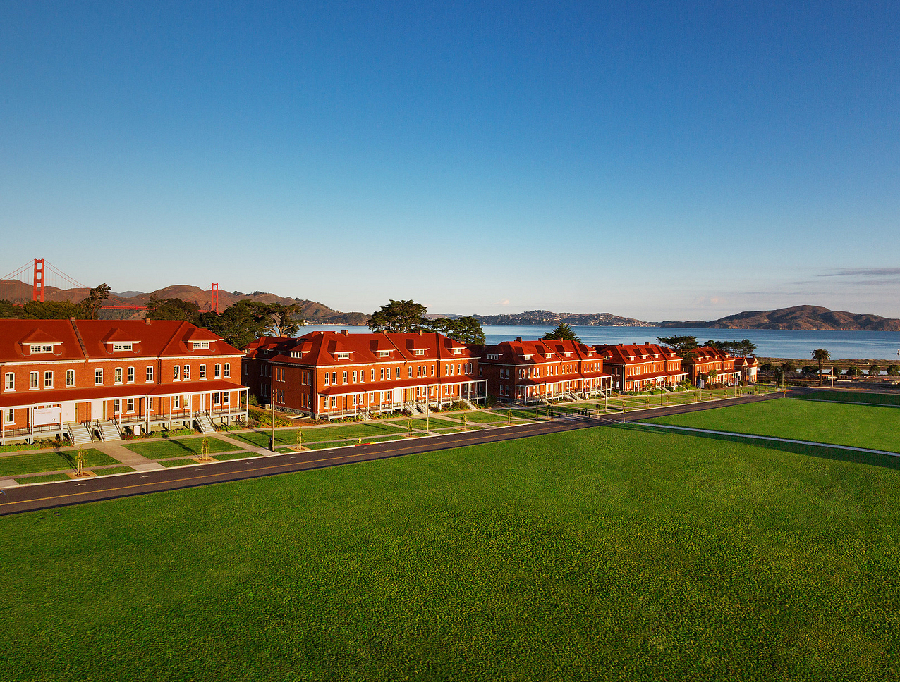 Row of red-roofed buildings with Golden Gate Bridge in the distance, beside a green lawn under clear blue skies.