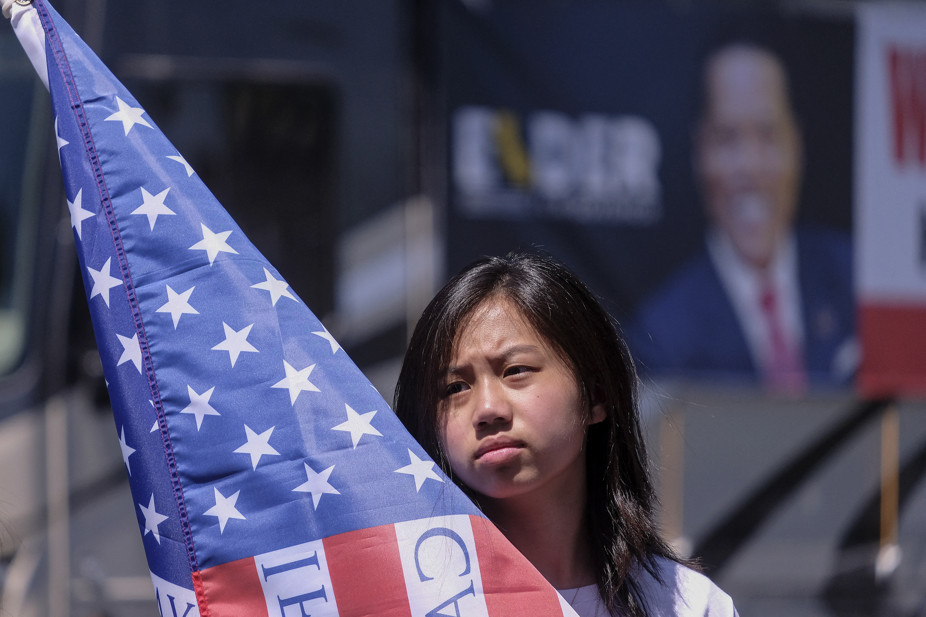A young woman stands in front of a blurred banner with an American flag partially obscuring her face.