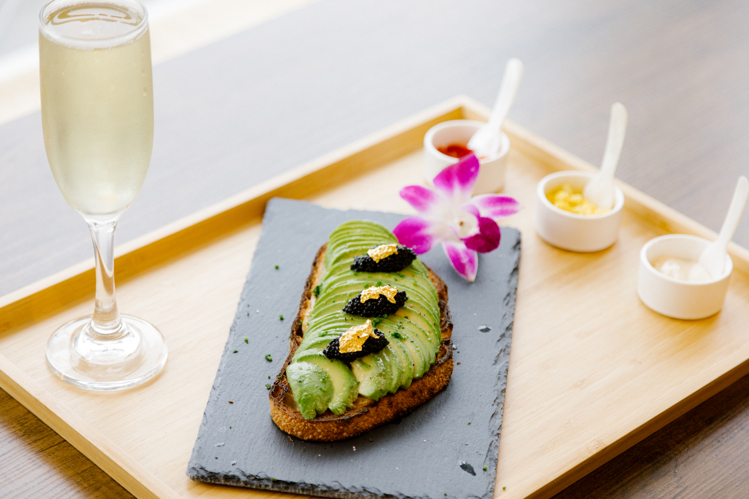 A slice of avocado toast with caviar and gold leaf on top sits on a tray with a glass of champagne and condiments.
