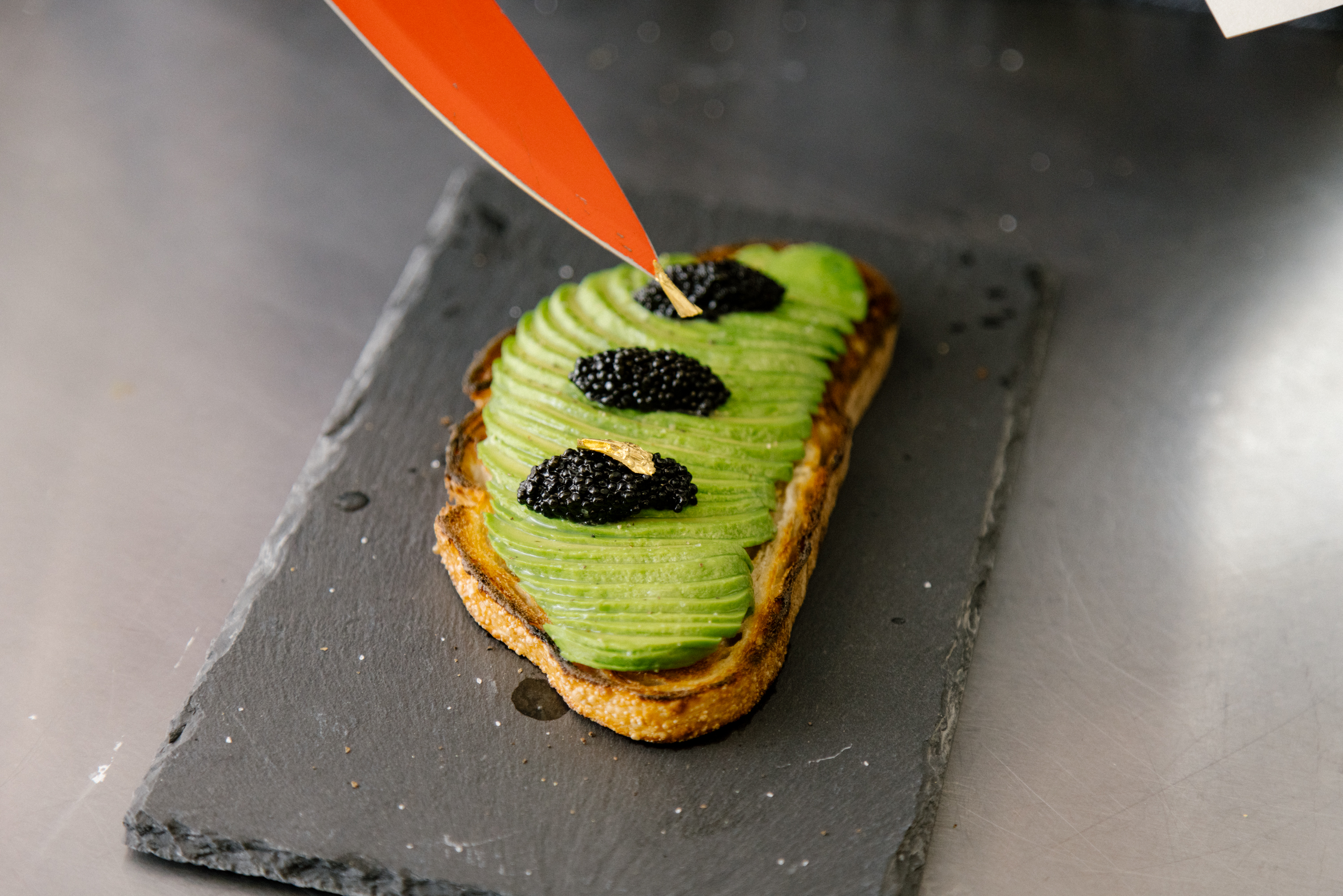 A slice of avocado toast topped with caviar and gold leaf sits atop a black slate slab.