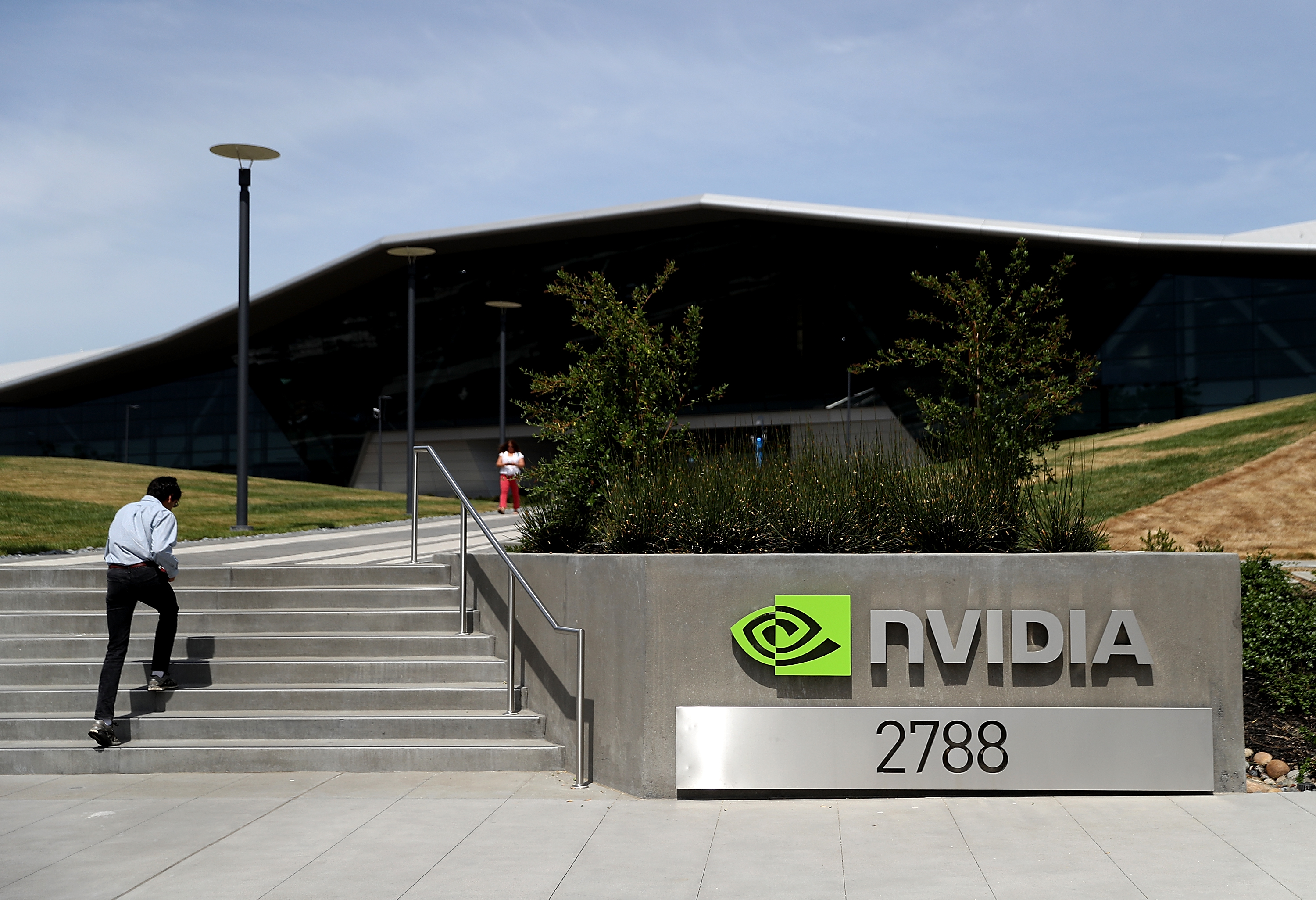 A man walks down steps at NVIDIA's modern office building entrance marked &quot;2788&quot; under a vibrant green logo. Another person stands in the background.
