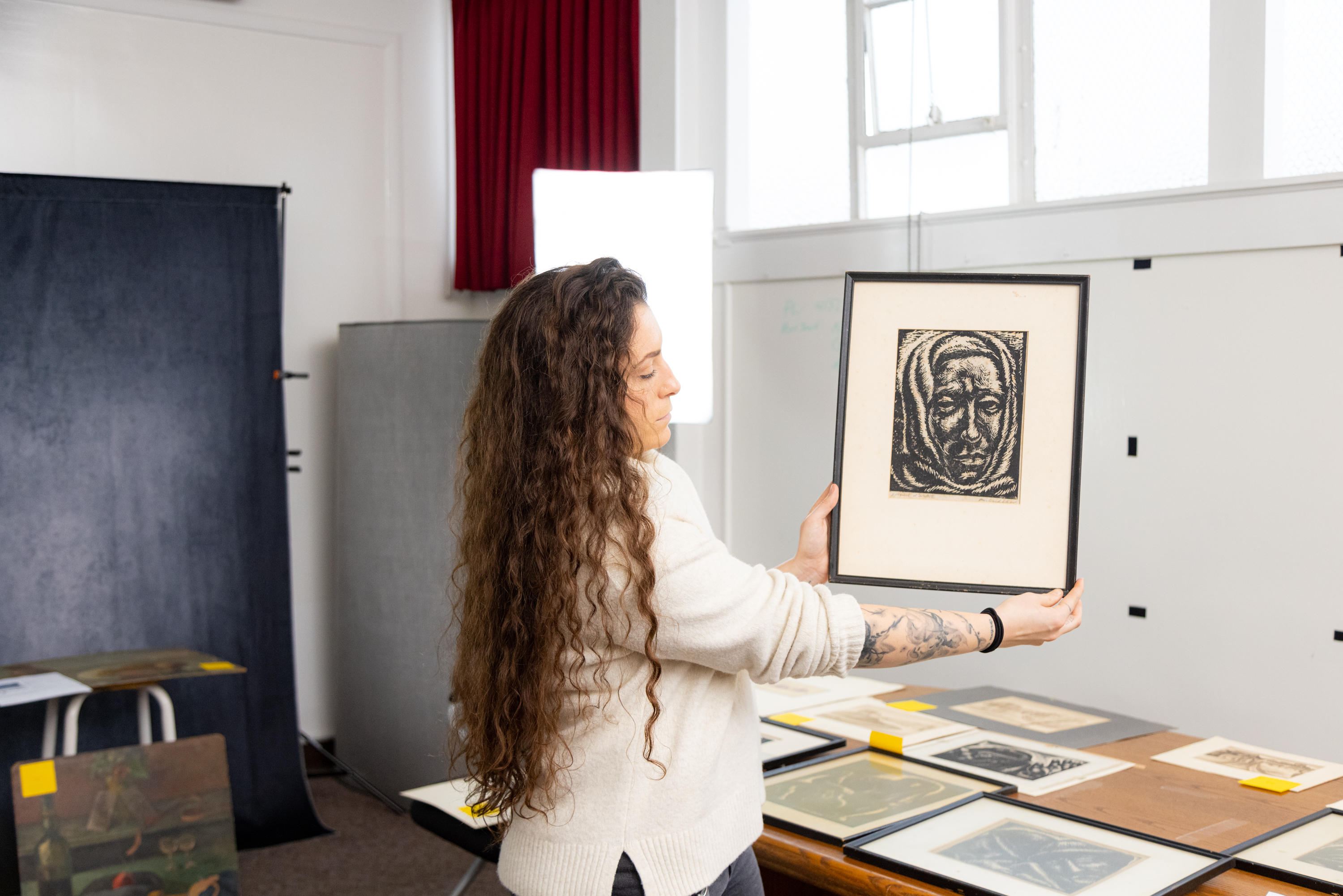A woman holding a framed print of a face, standing near a table with more artwork.