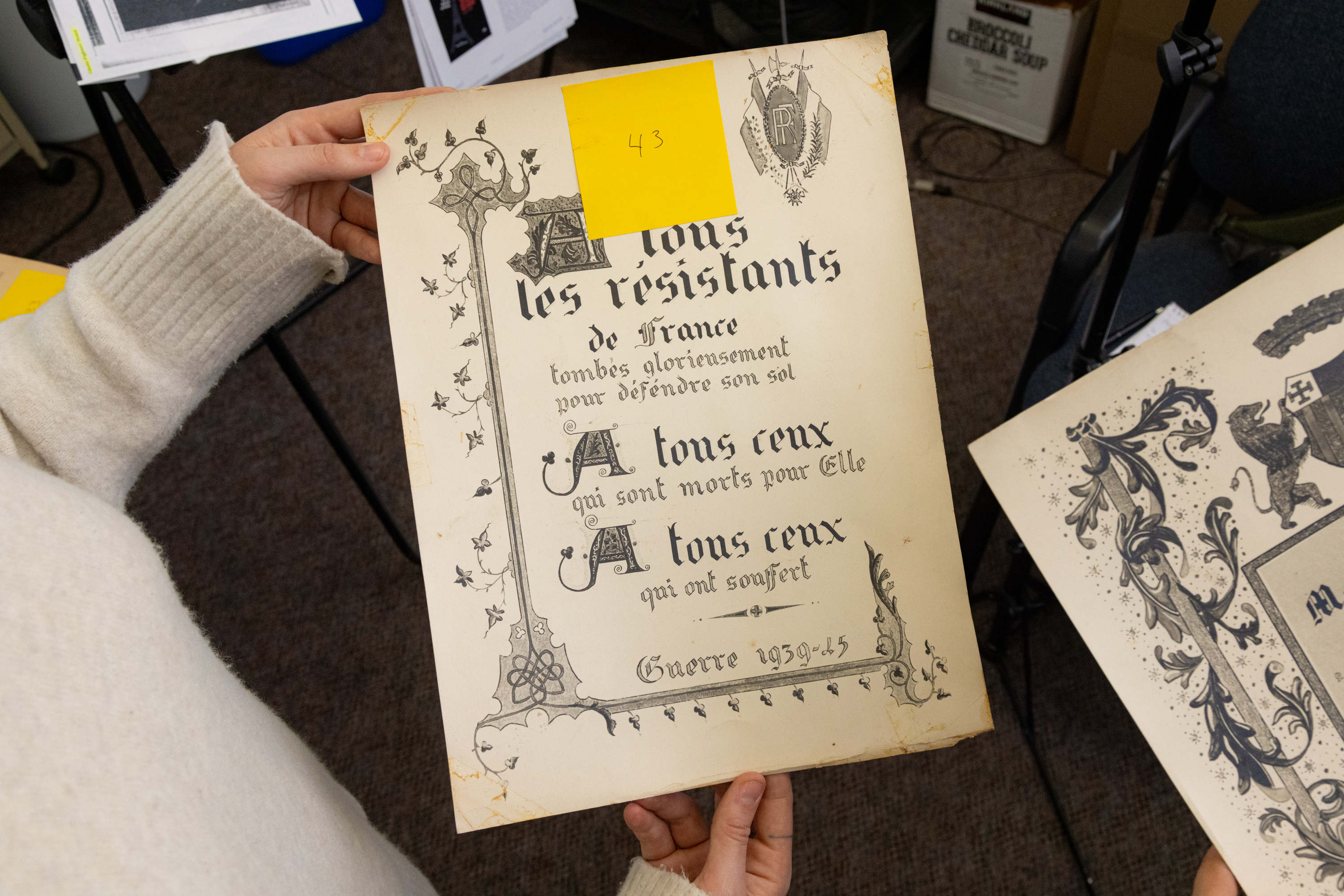 Hands holding an old document with French text, floral designs, and a yellow sticky note.