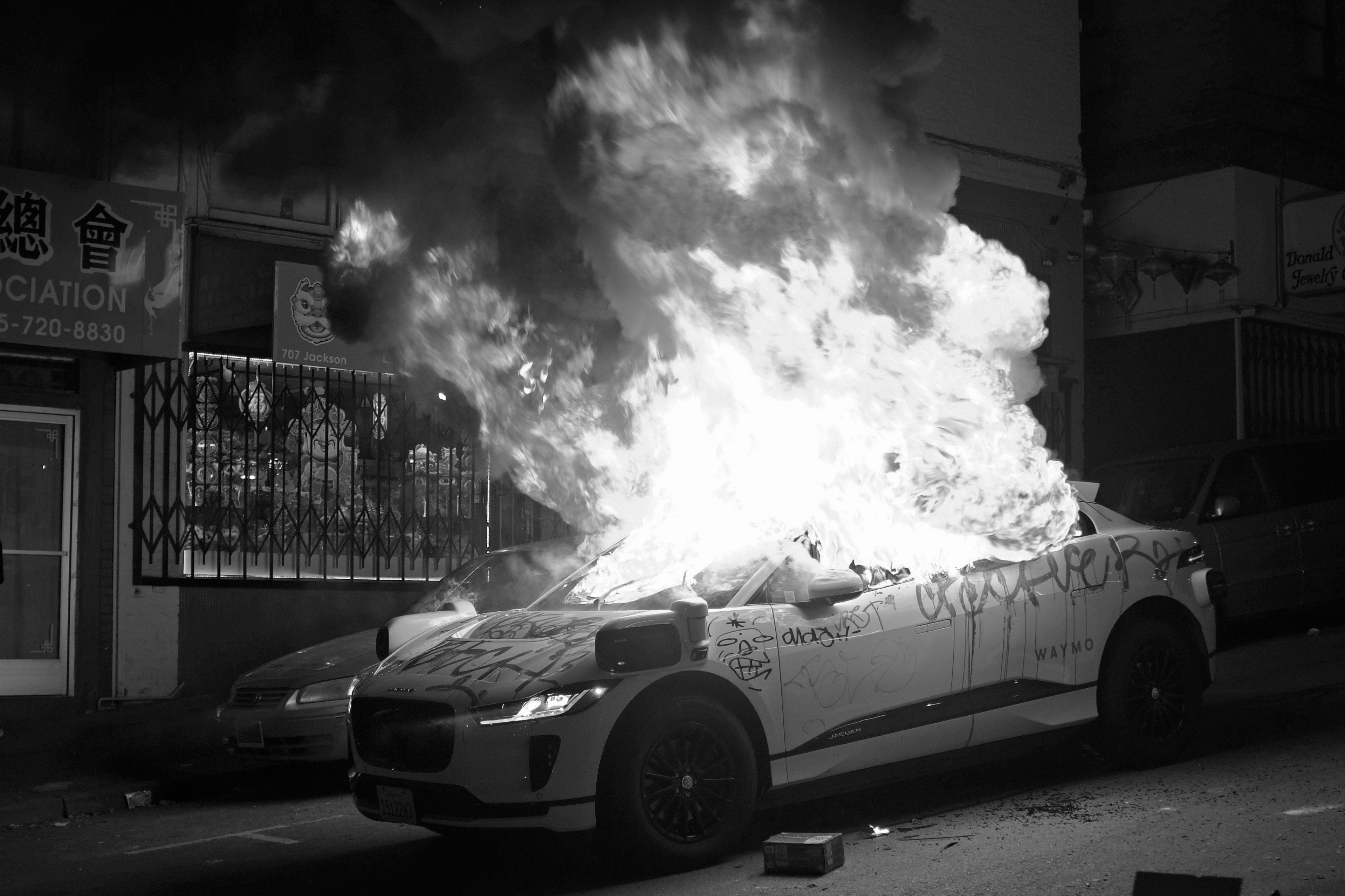 A car engulfed in flames on a city street at night, with graffiti on its side.