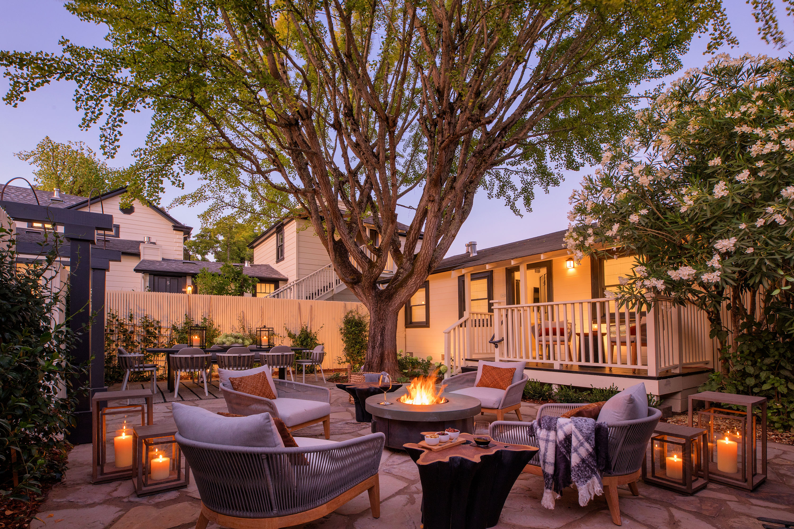 Wicker chairs with cushions sit around a fireplace under a large tree in a backyard patio