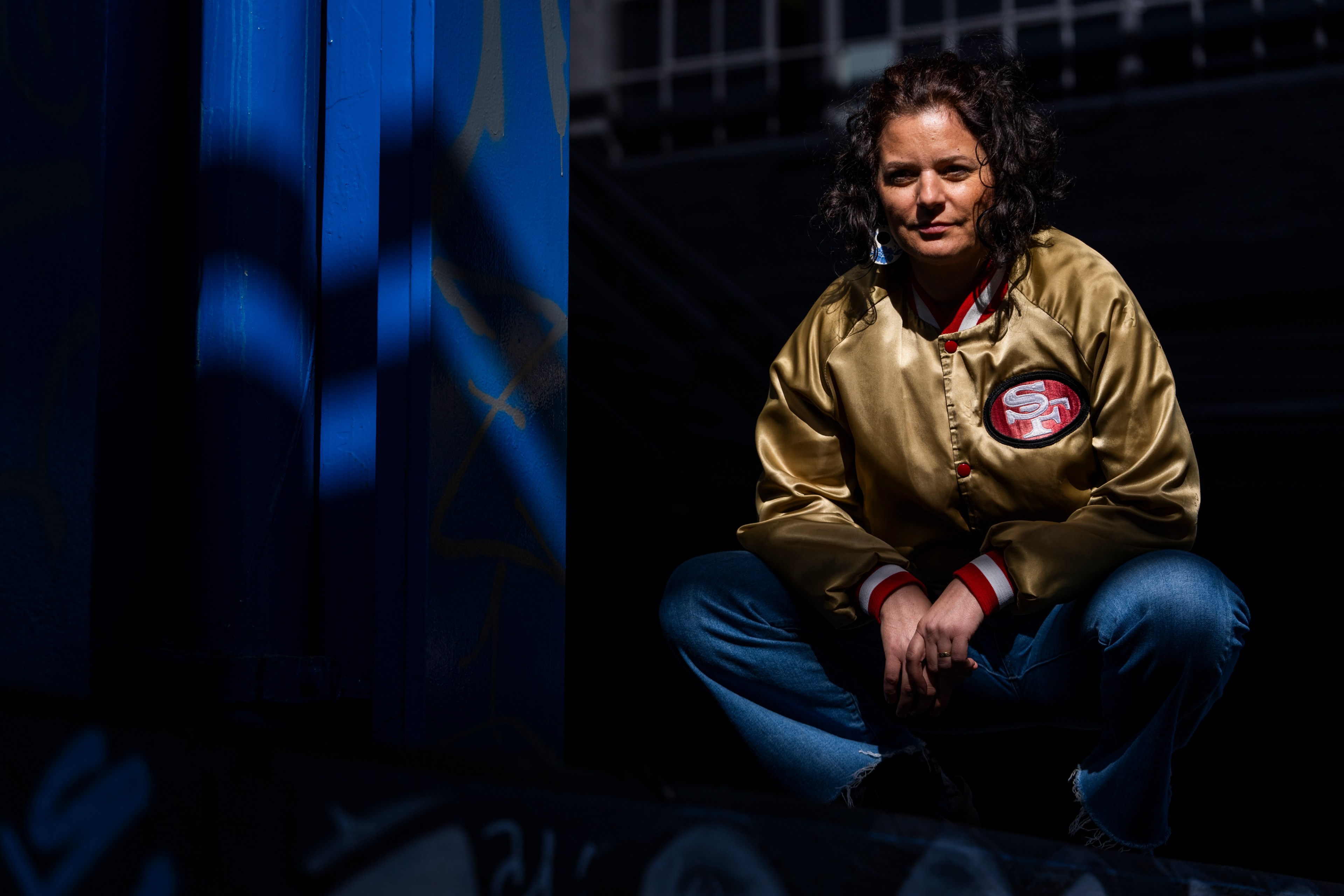 A person in a gold and red sports jacket squats in a shadowy area with graffiti, sunlit face and jacket prominent.